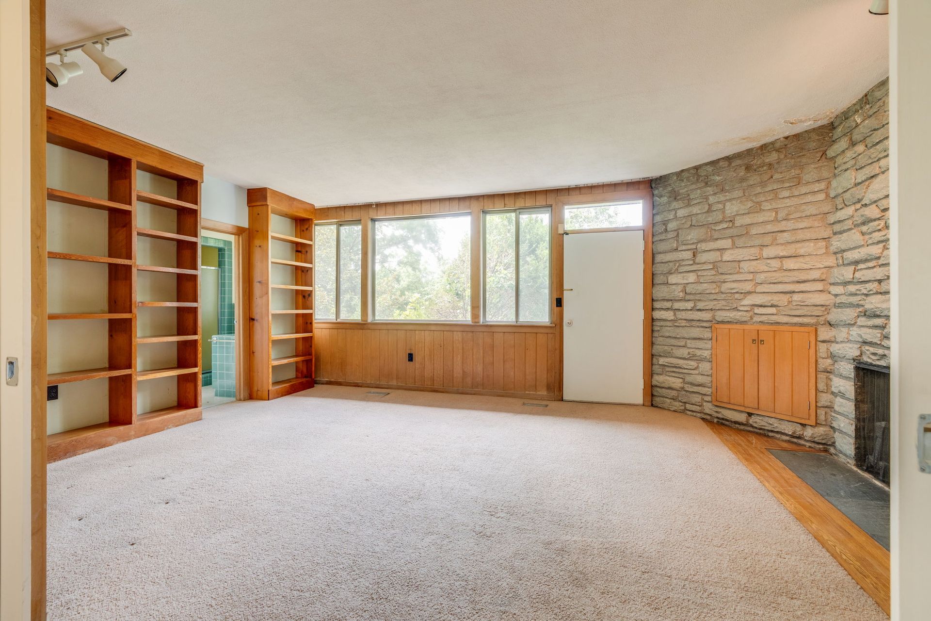 Empty living room with stone accent wall, large windows, built-in shelves, and carpeted floor.