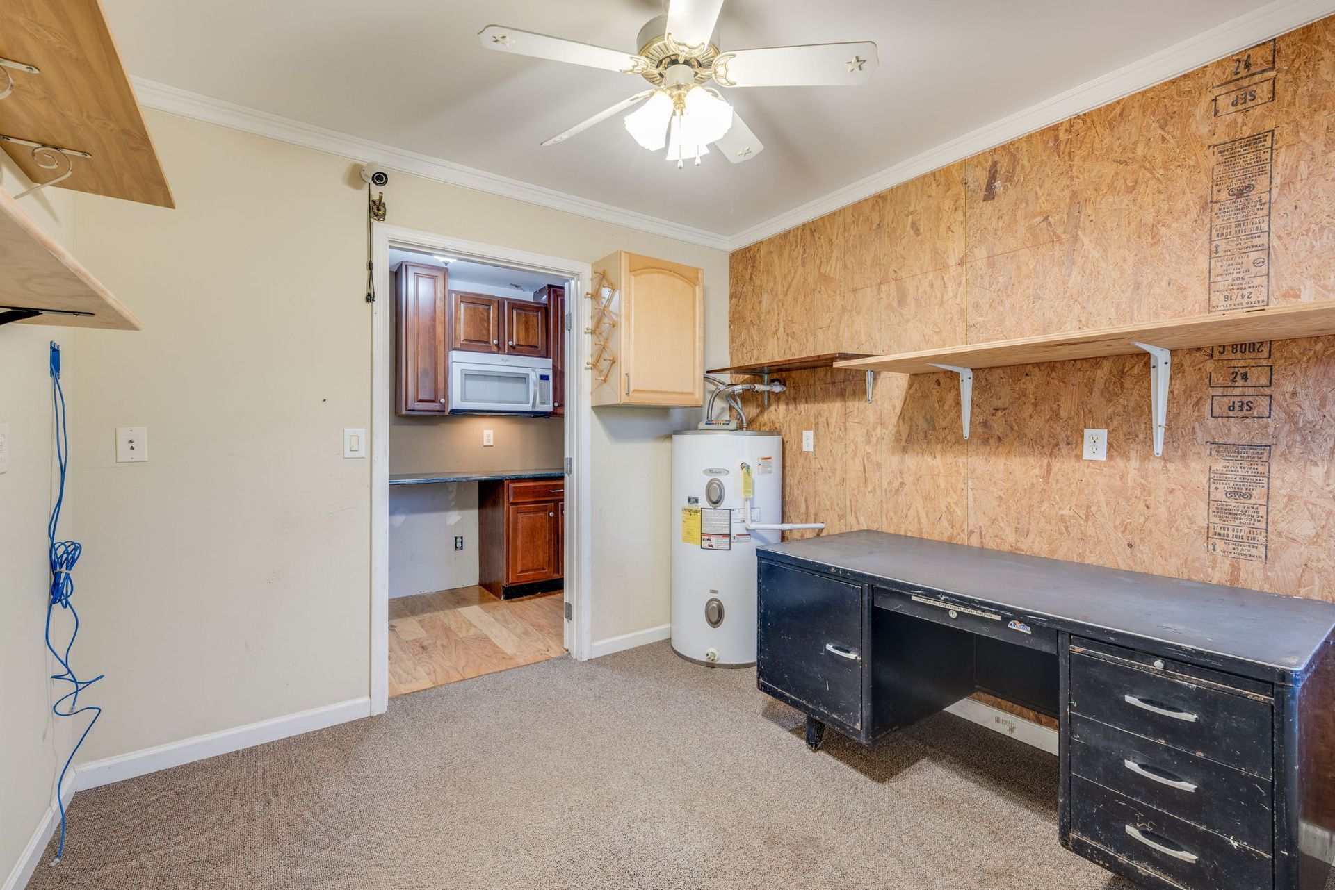 Small room with desk, shelving, ceiling fan, and doorway to a kitchen area