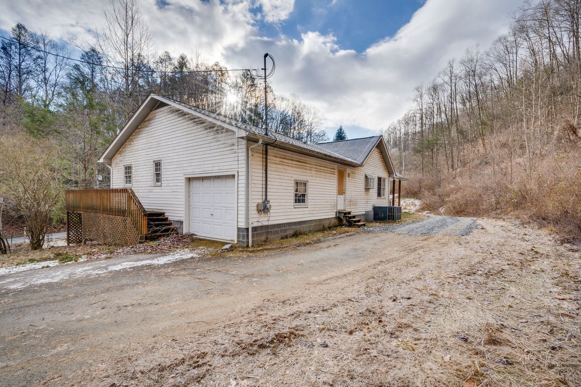 White house with a garage and wooden deck, on a dirt road in a wooded area. Cloudy sky.