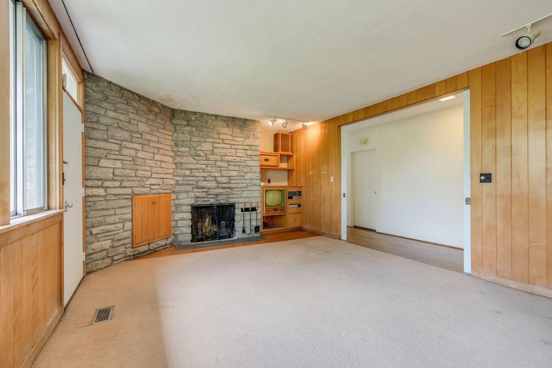 Empty living room with stone fireplace, wood-paneled walls, and beige carpet.
