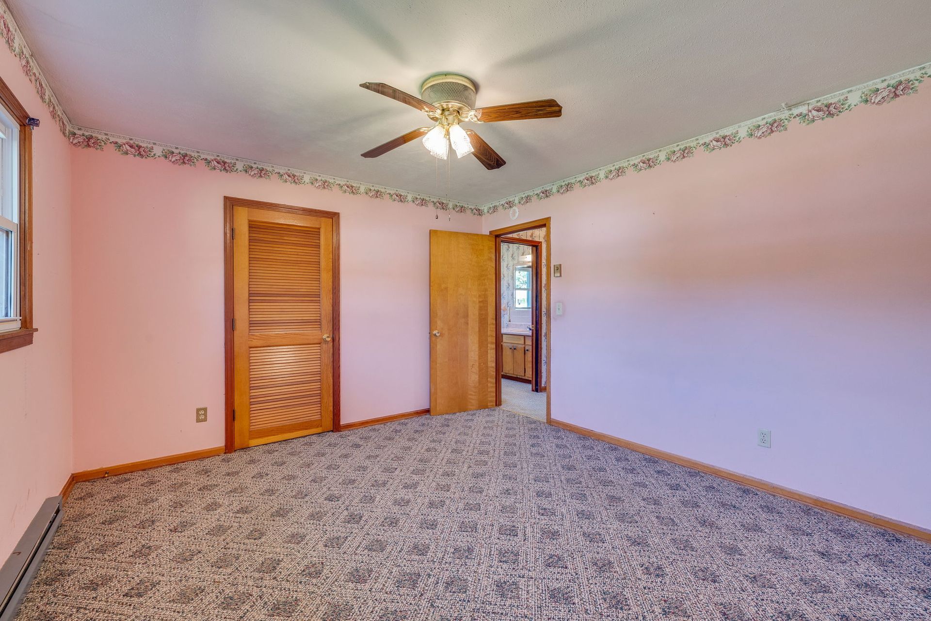 Empty bedroom with pink walls, patterned carpet, ceiling fan, and wooden closet doors.