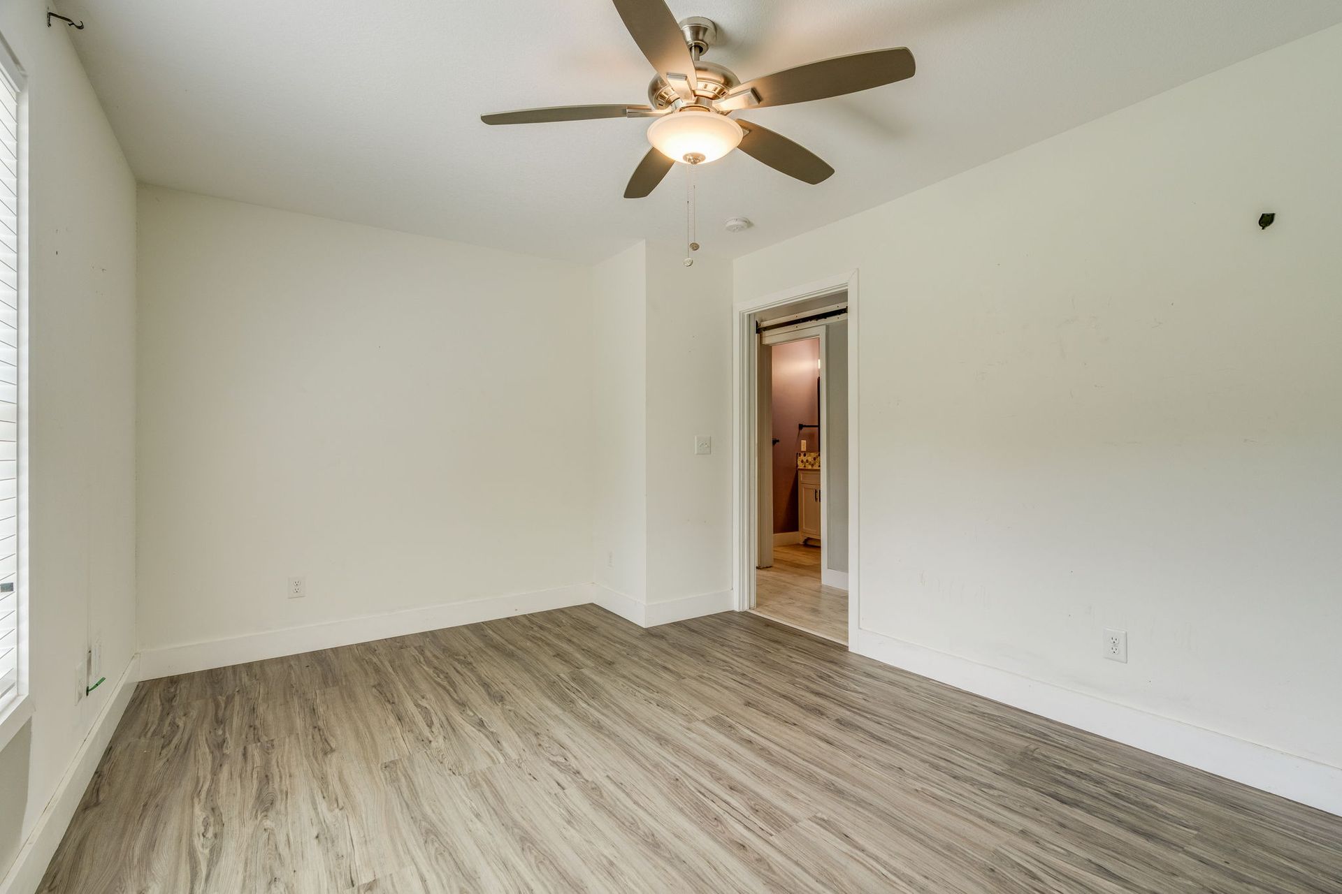 Empty bedroom with white walls, wood floor, ceiling fan, and open doorway to a bathroom