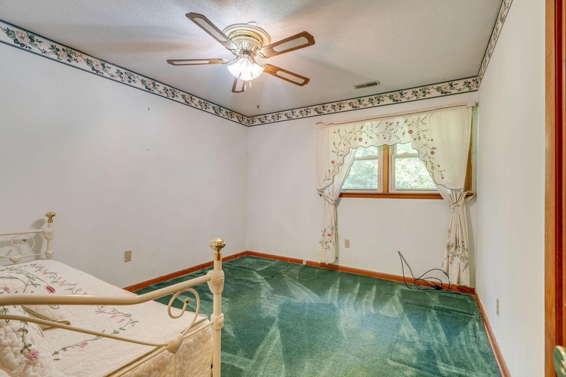 Empty bedroom with green carpet, brass bed frame, ceiling fan, and window with lace curtains