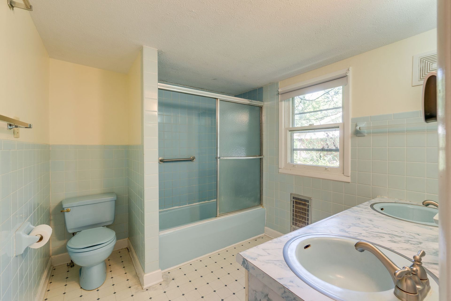 Small bathroom with blue shower, toilet, and sink, white tiled floor, and window.