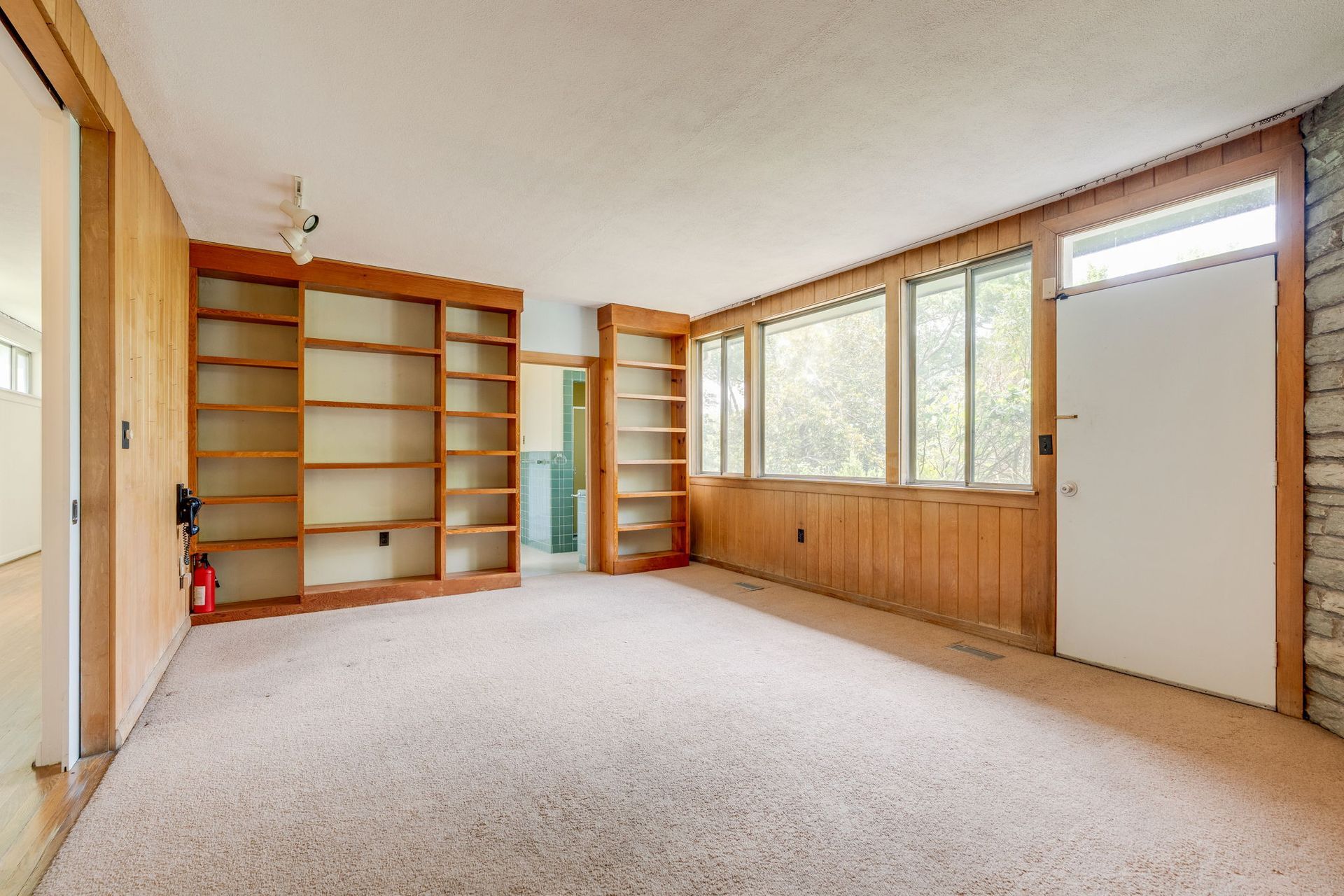 Empty carpeted room with built-in wooden shelves and large windows.