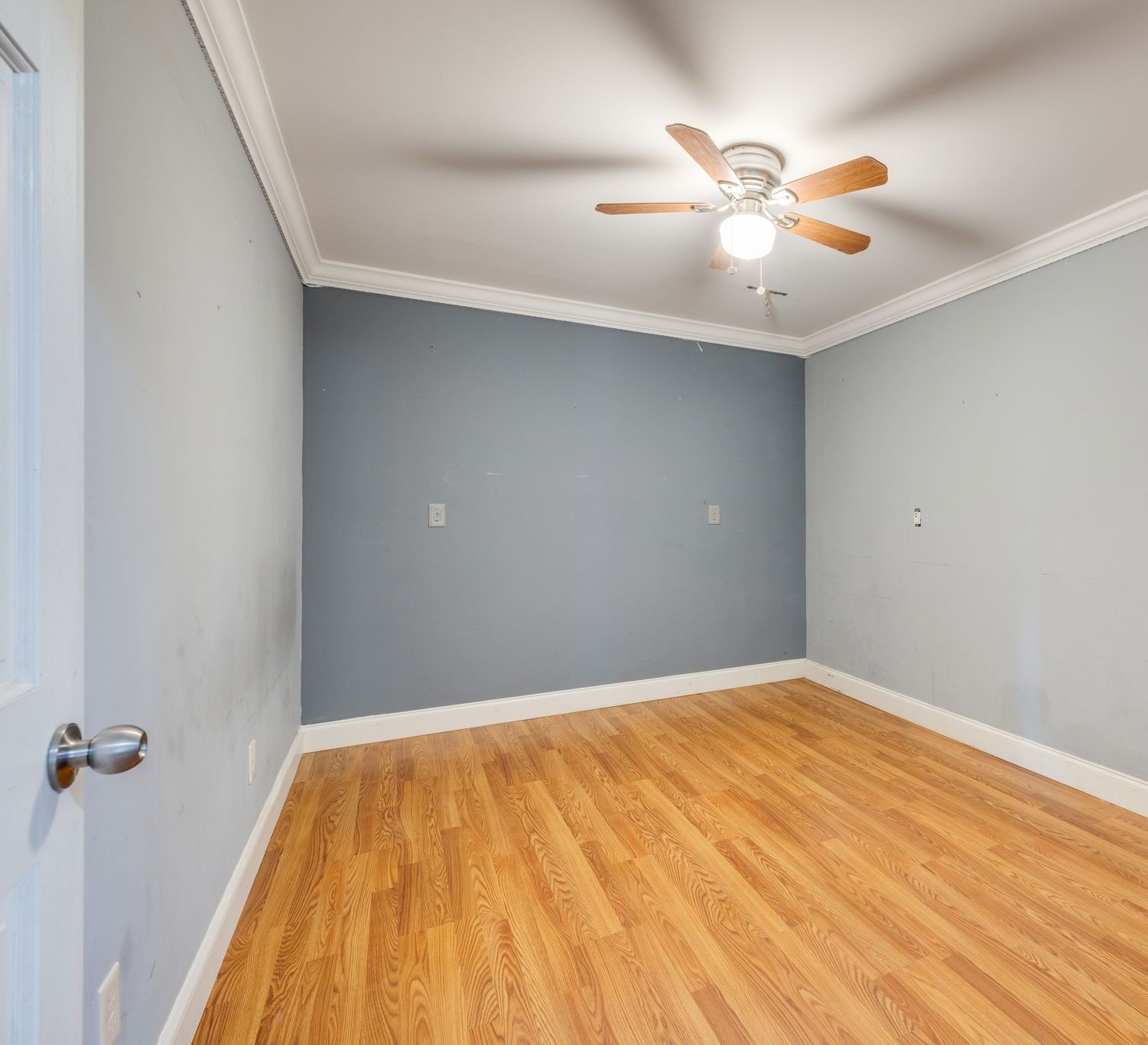 Empty bedroom with gray walls, hardwood floor, white trim, and a ceiling fan with lights.