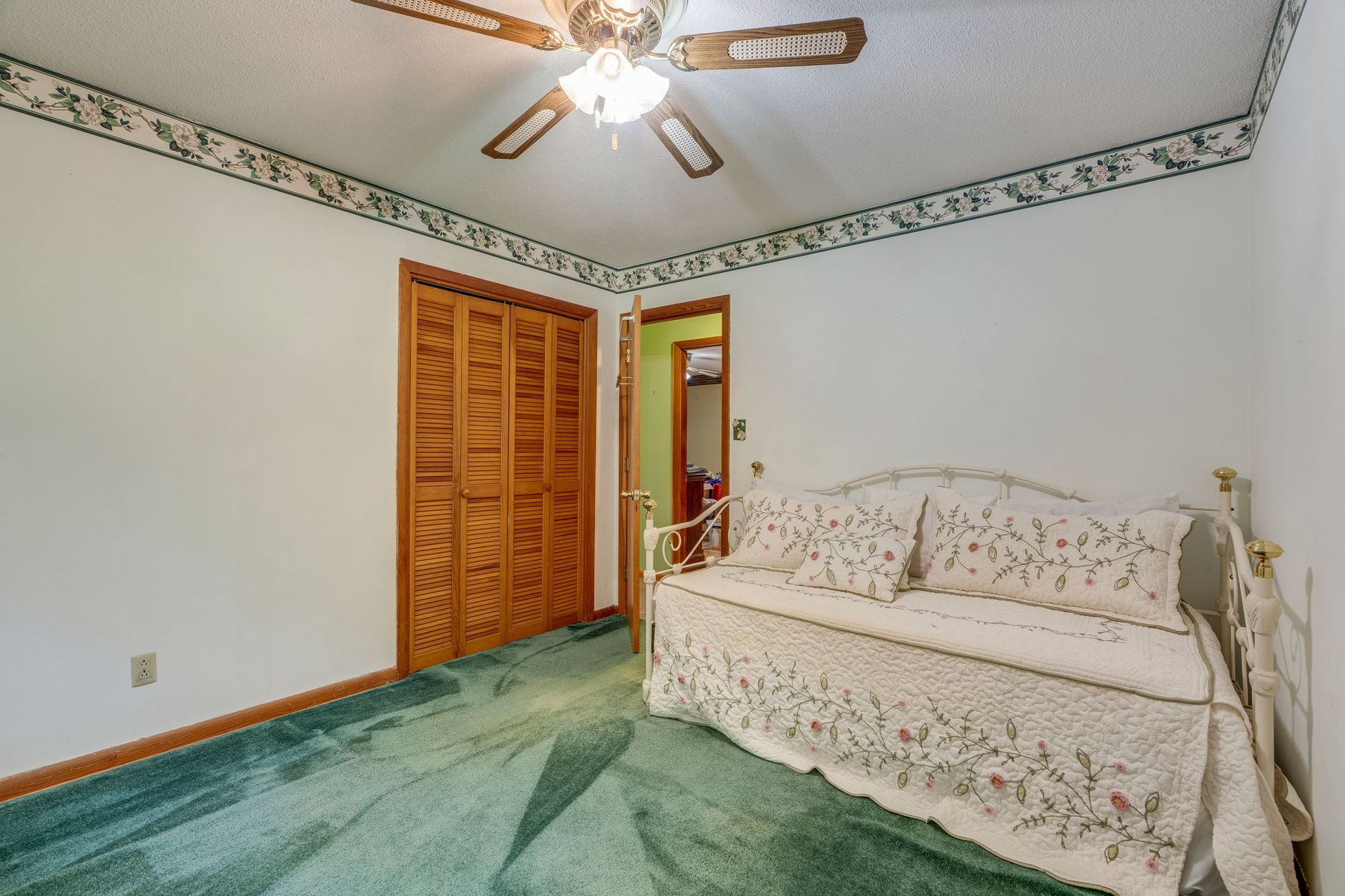 Bright bedroom with green carpet, white floral couch, wood closet doors, and ceiling fan.