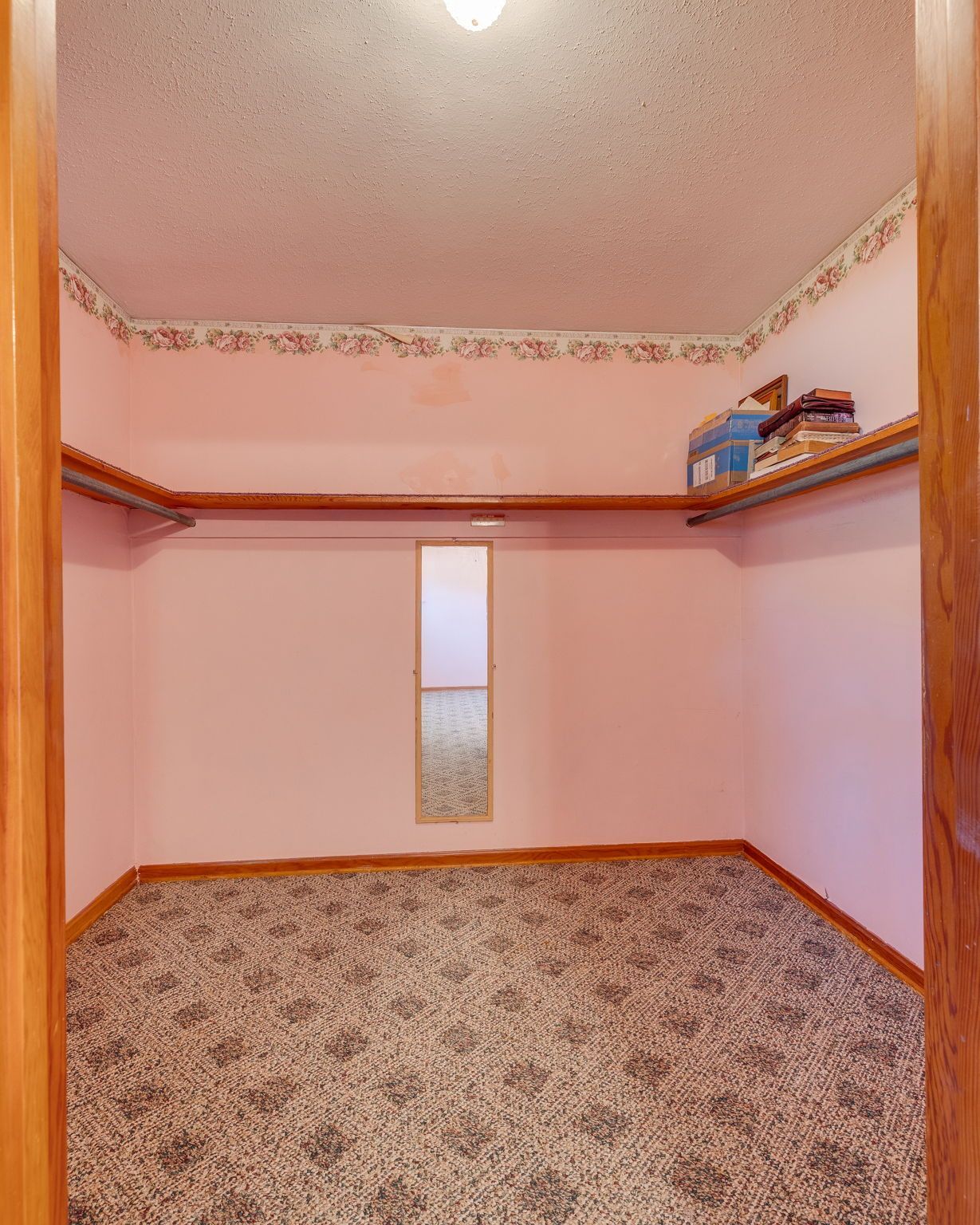Empty pink walk-in closet with patterned carpet, wood trim, and a shelf holding folded items.