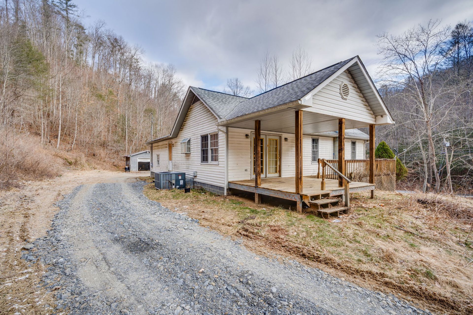 White house with porch and gravel driveway in a wooded, hilly setting under a cloudy sky.