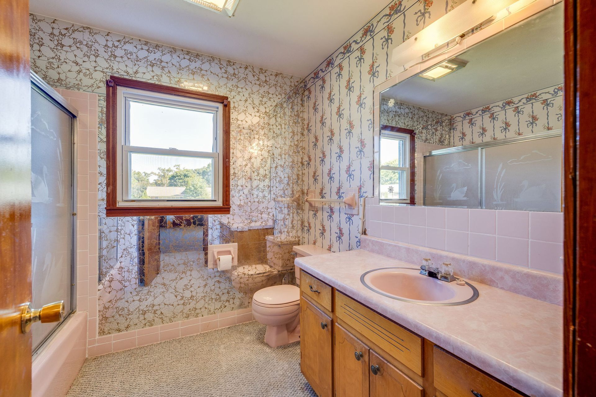 Bathroom with floral wallpaper, white tile, wooden vanity, sink, mirror, and window.