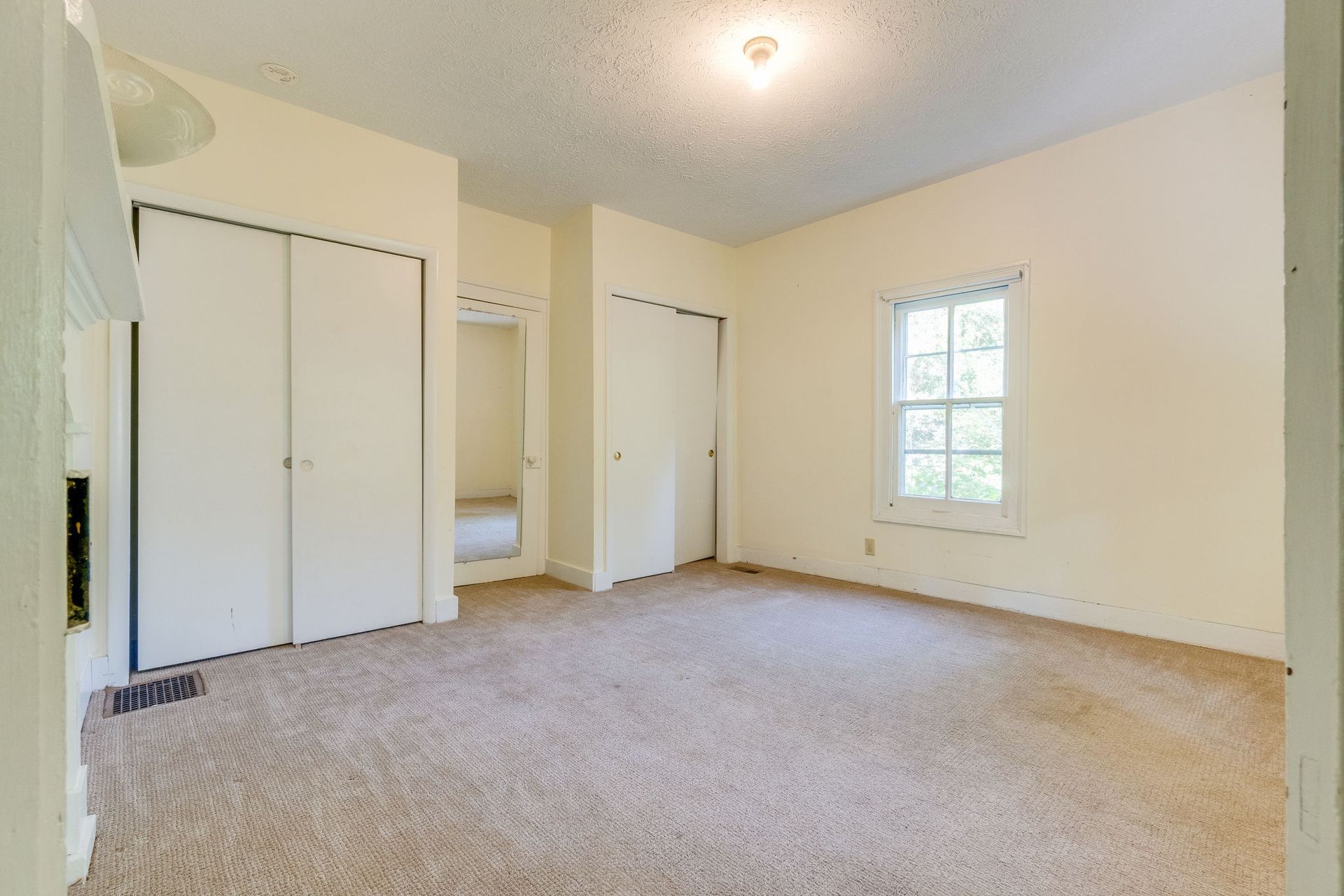 Empty beige bedroom with carpet, white closet doors, and a window on the right.