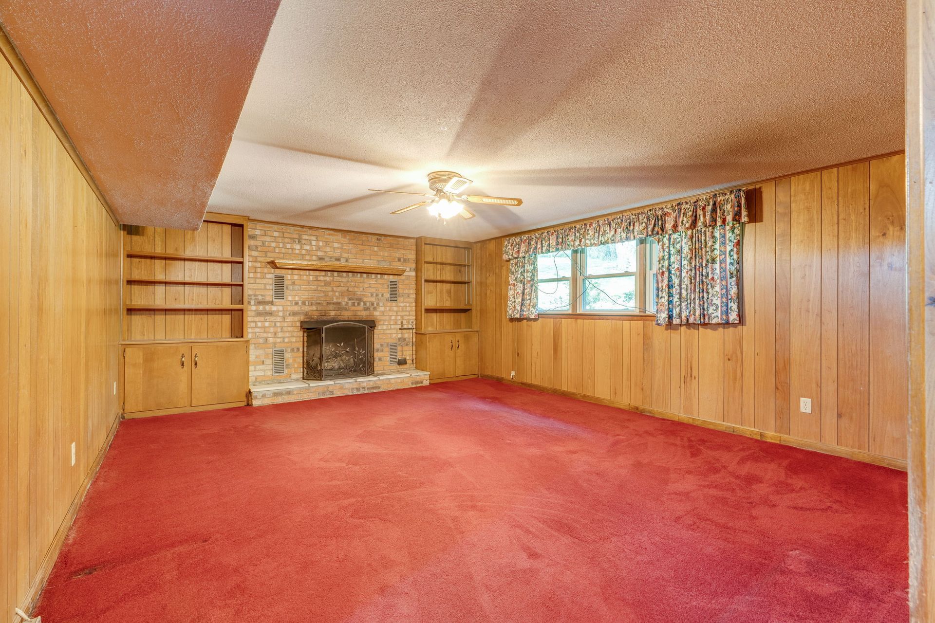 Empty wood-paneled living room with red carpet, brick fireplace, ceiling fan, and window curtains