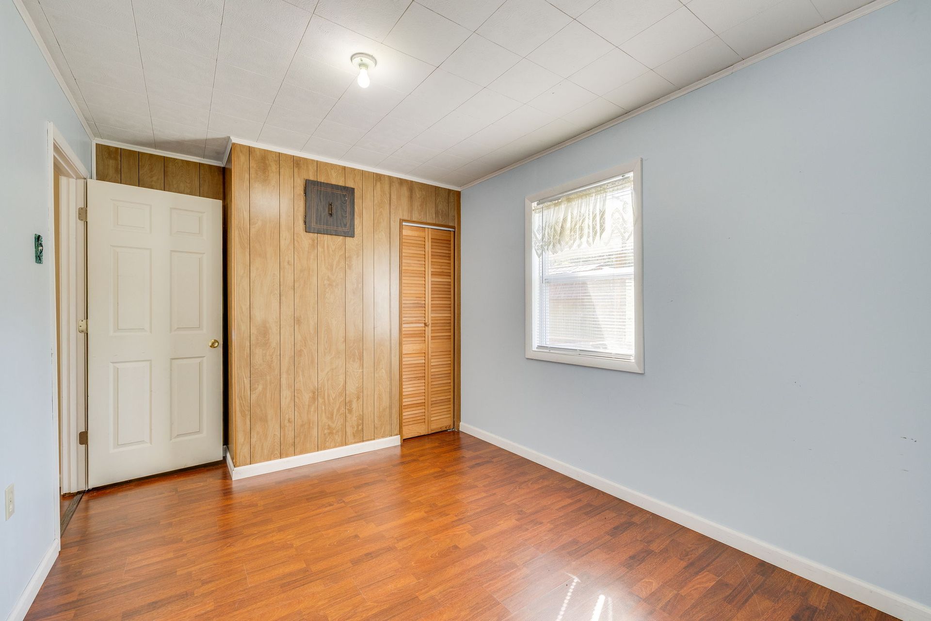 Empty room with hardwood floor, white walls, wood-paneled accent wall, closet door, and a window.
