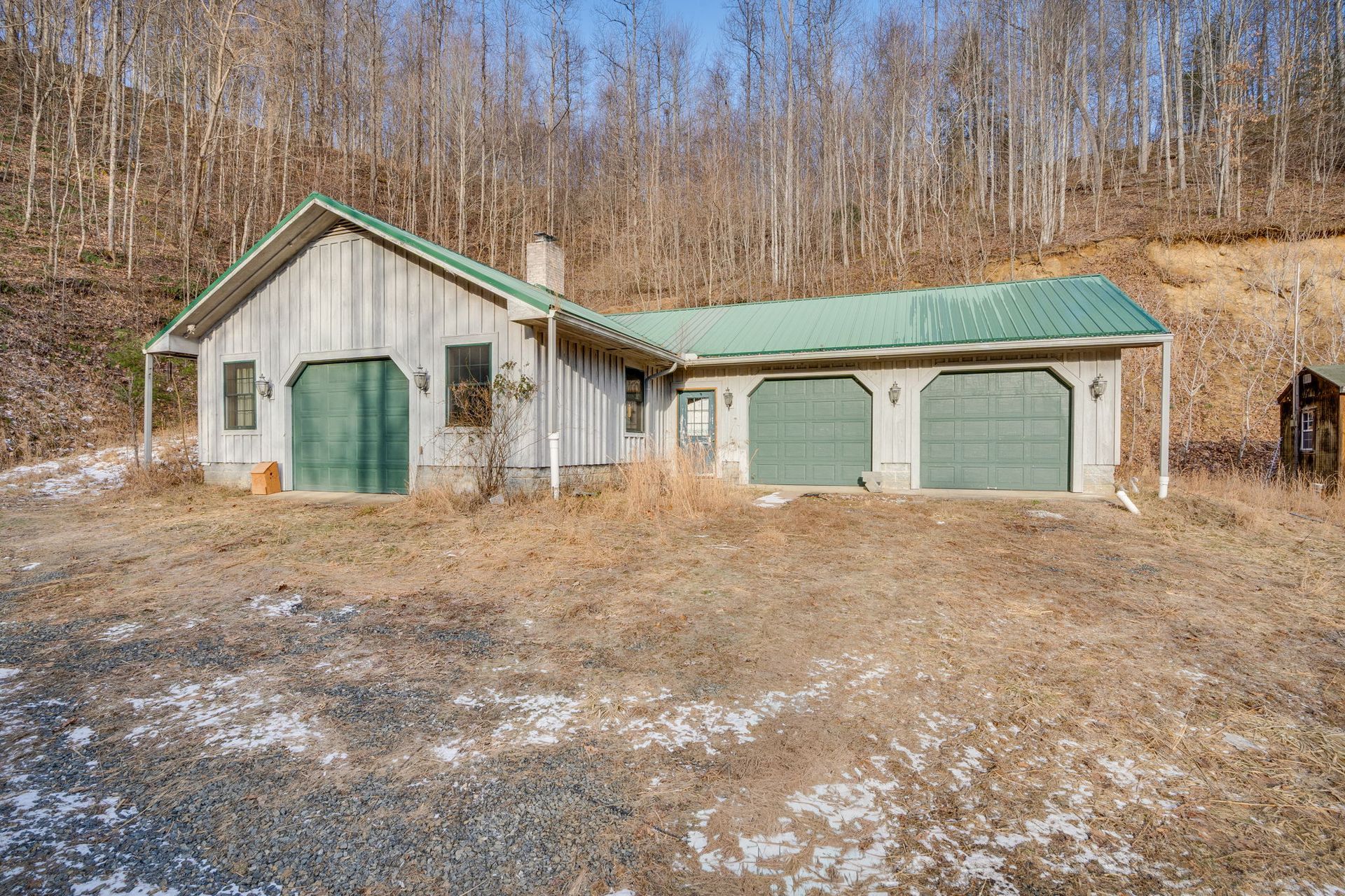 Rustic garage with three green doors, a green roof, and a chimney, set in a dry, grassy landscape with trees.