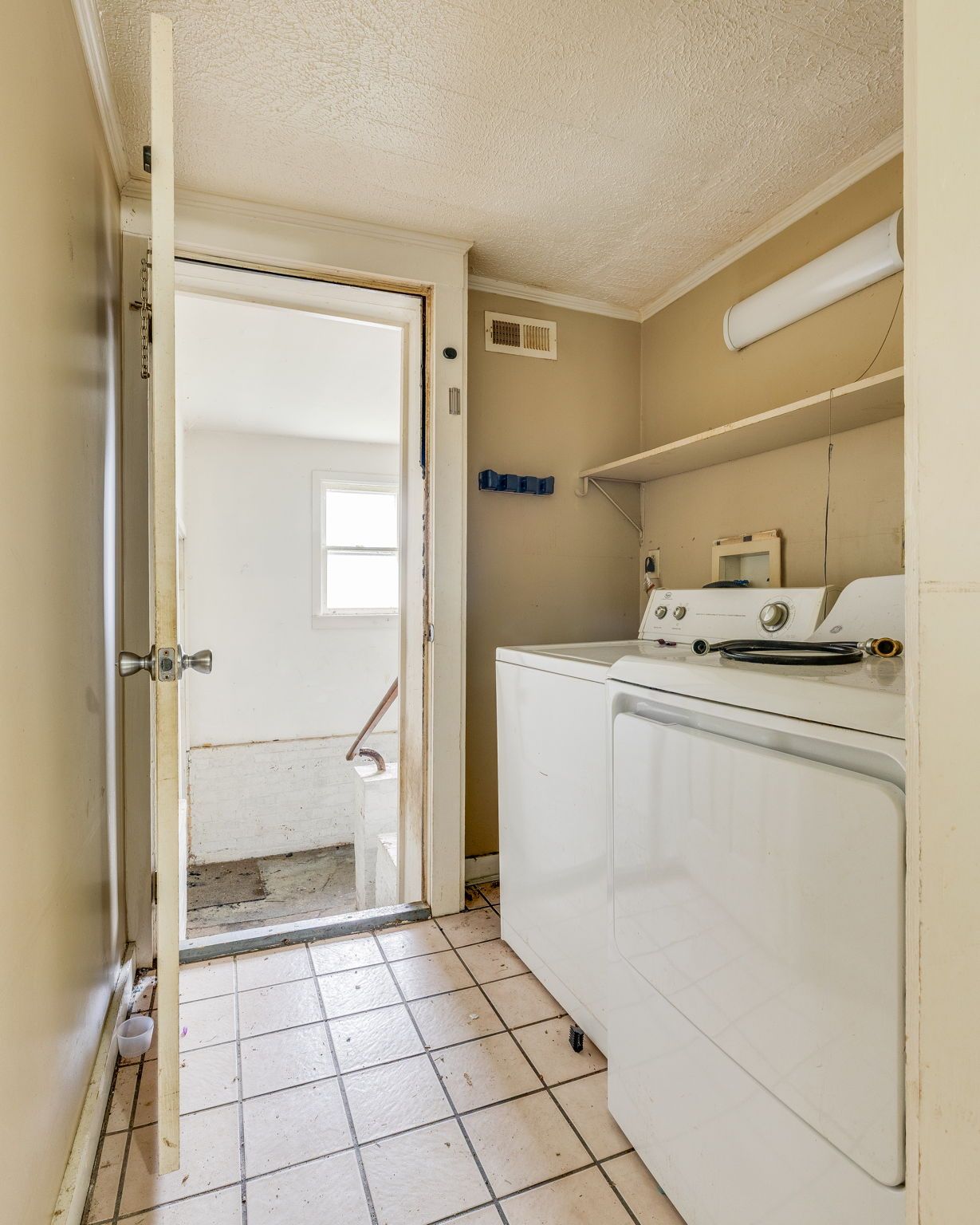 Small beige laundry room with washer, shelves, and open door to a bright utility room