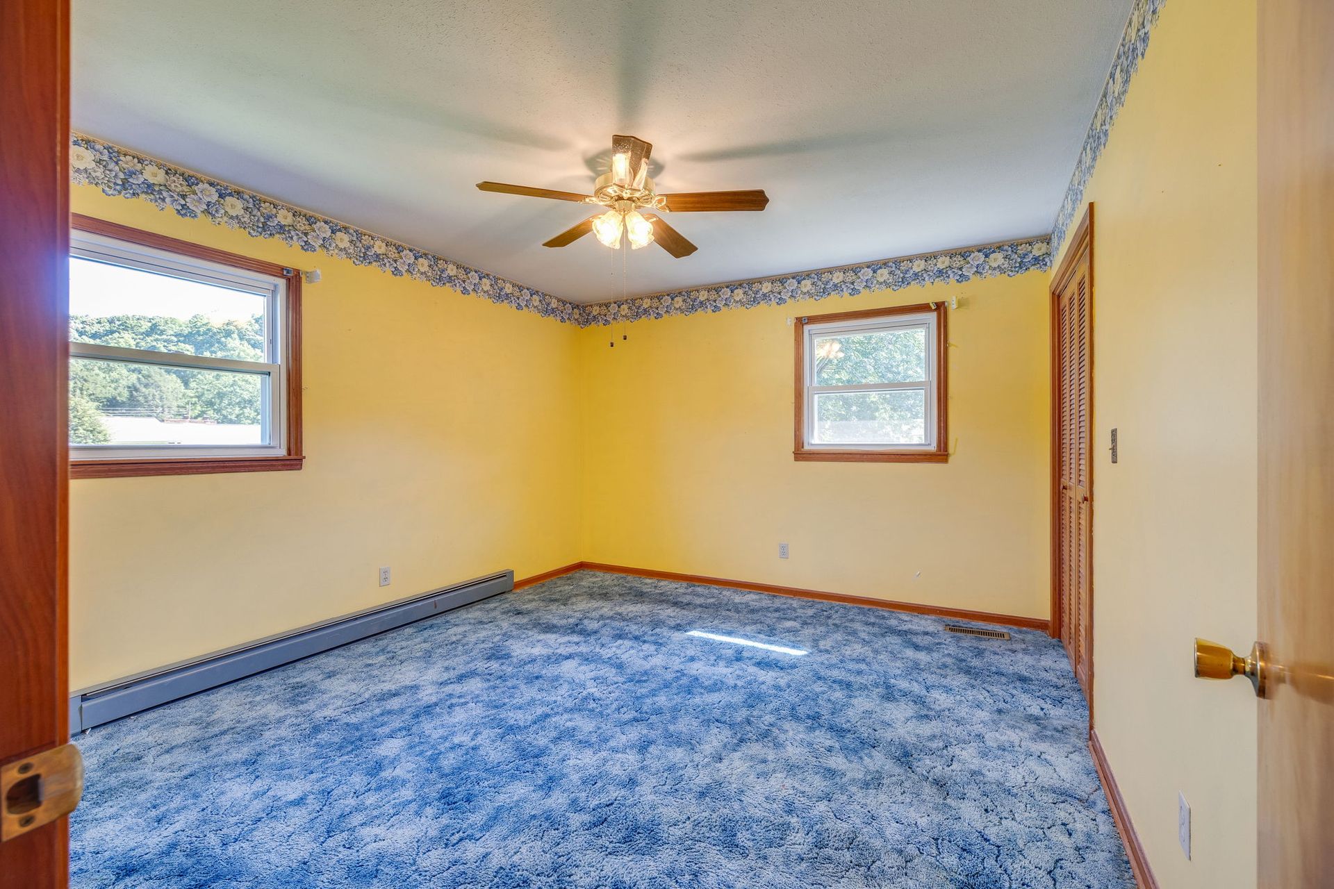 Empty yellow bedroom with blue carpet, ceiling fan, and two windows.