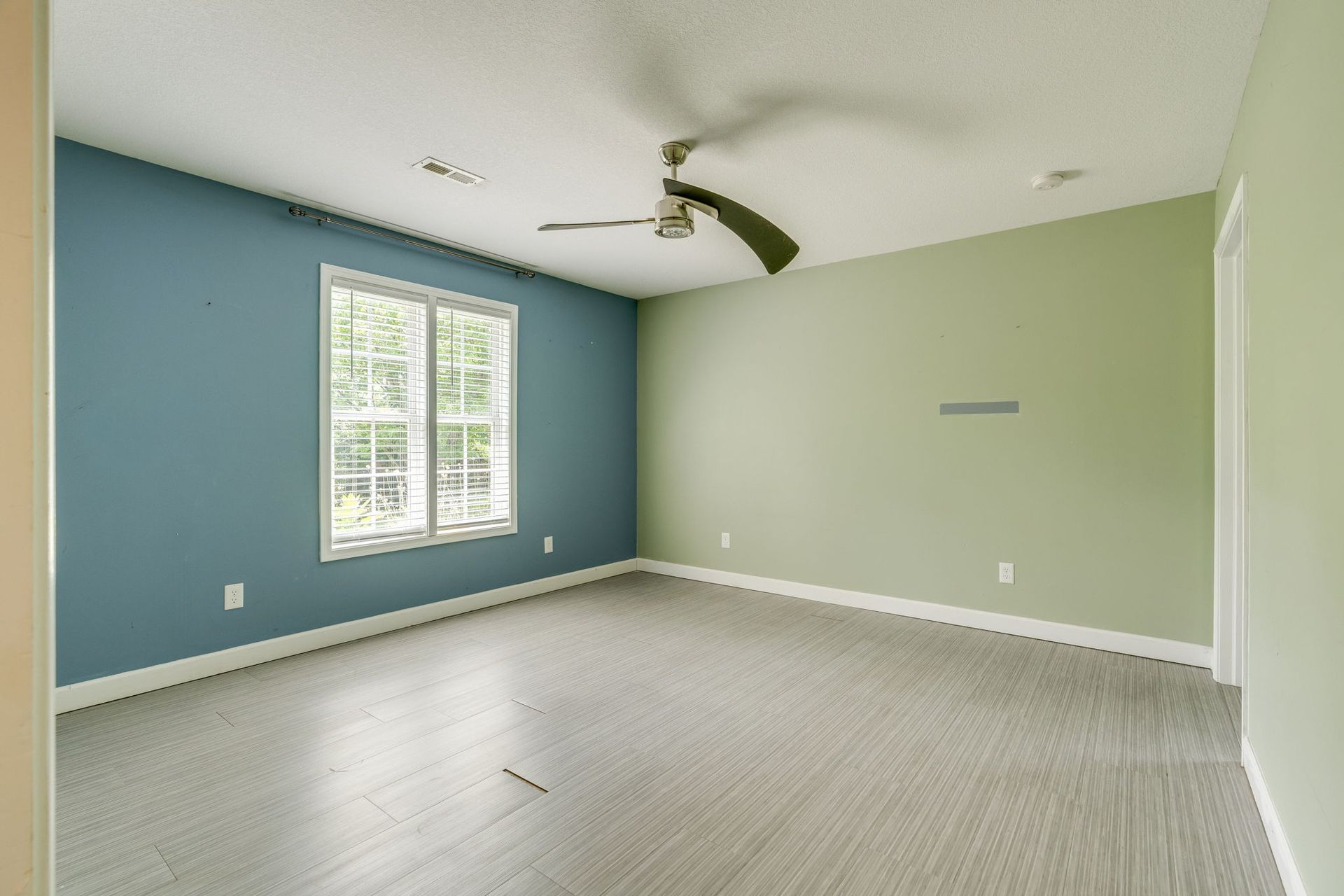 Empty bedroom with blue and green walls, white carpet, and a ceiling fan near a window.