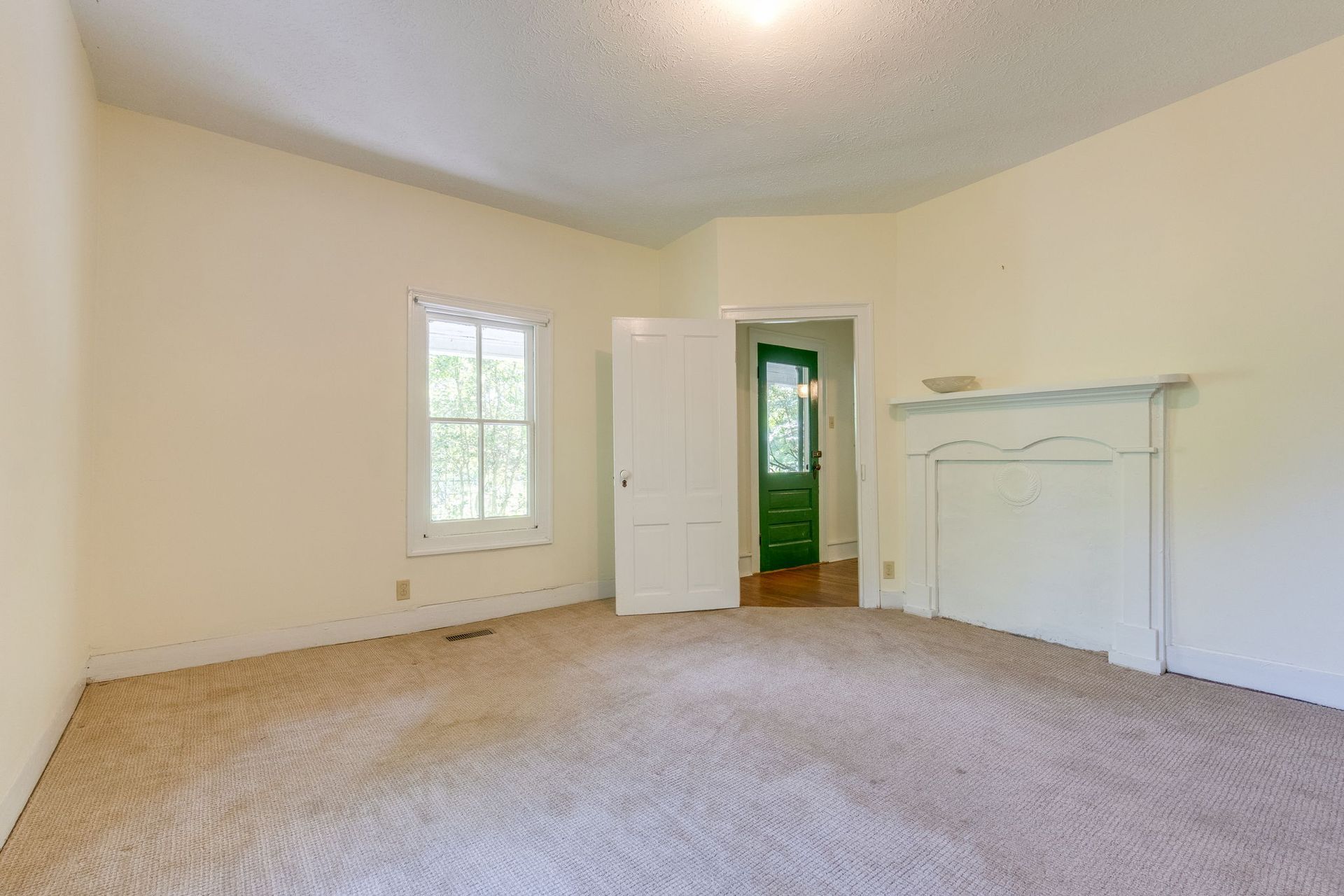 Empty beige bedroom with carpet, window, open door, and white fireplace mantle