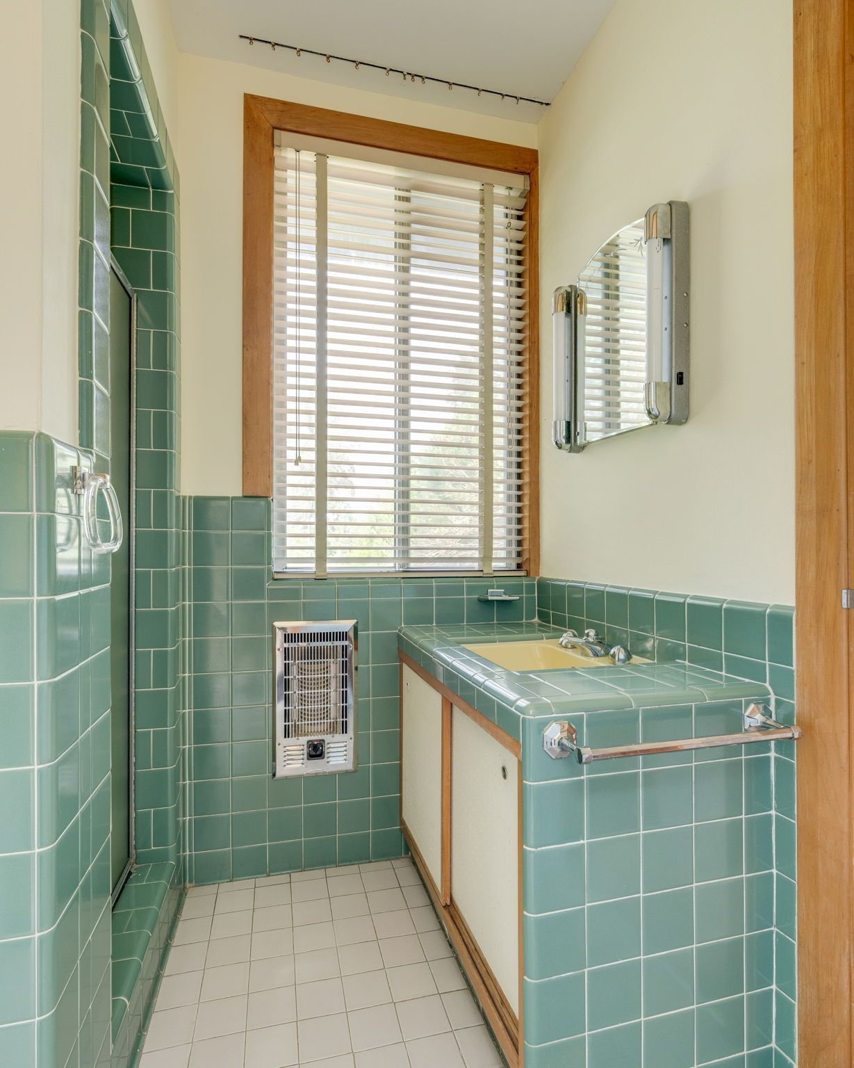 Narrow bathroom with green tile, pedestal sink, wall mirror, and window with wooden blinds.