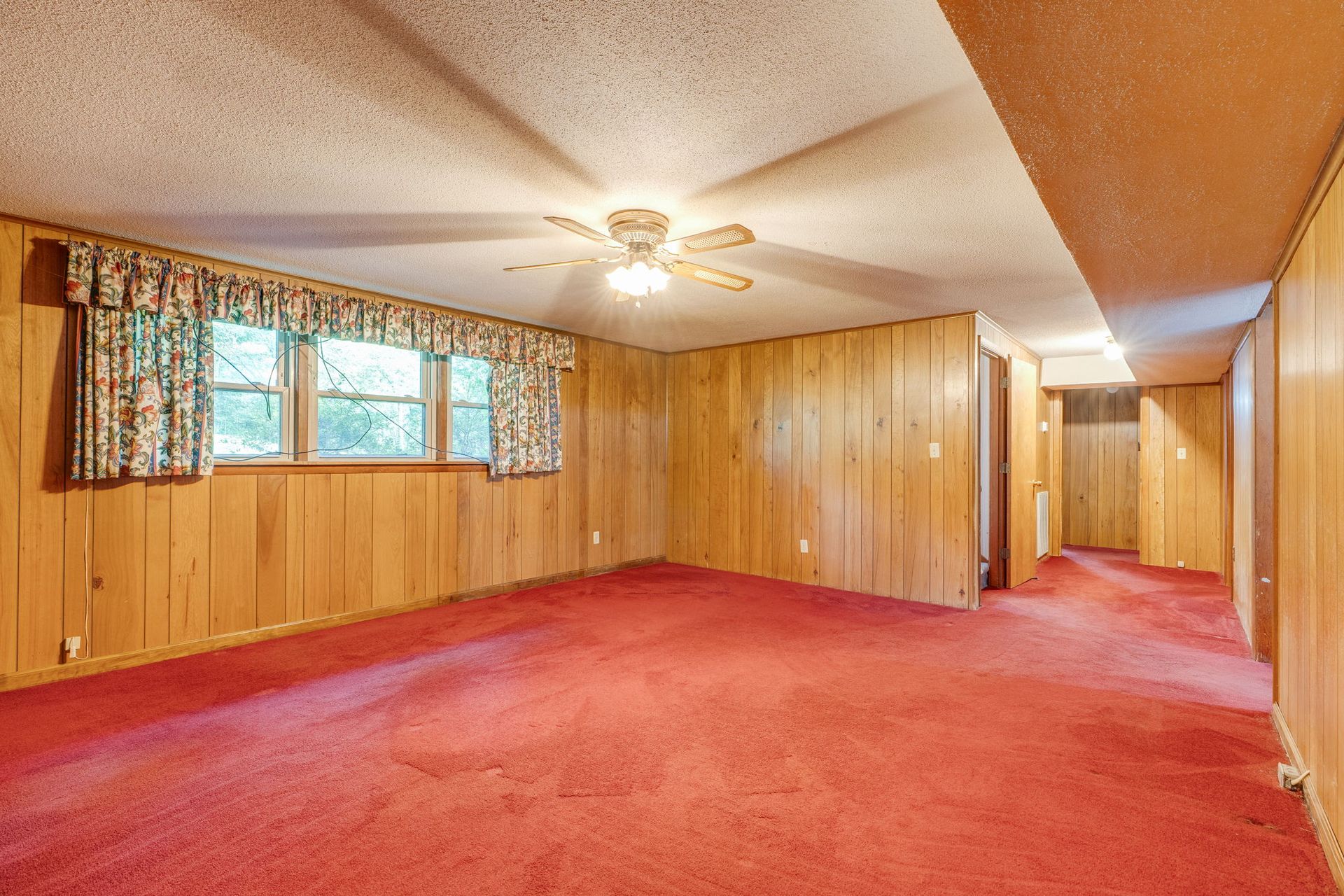 Empty wood-paneled living room with red carpet, window, ceiling fan, and hallway opening