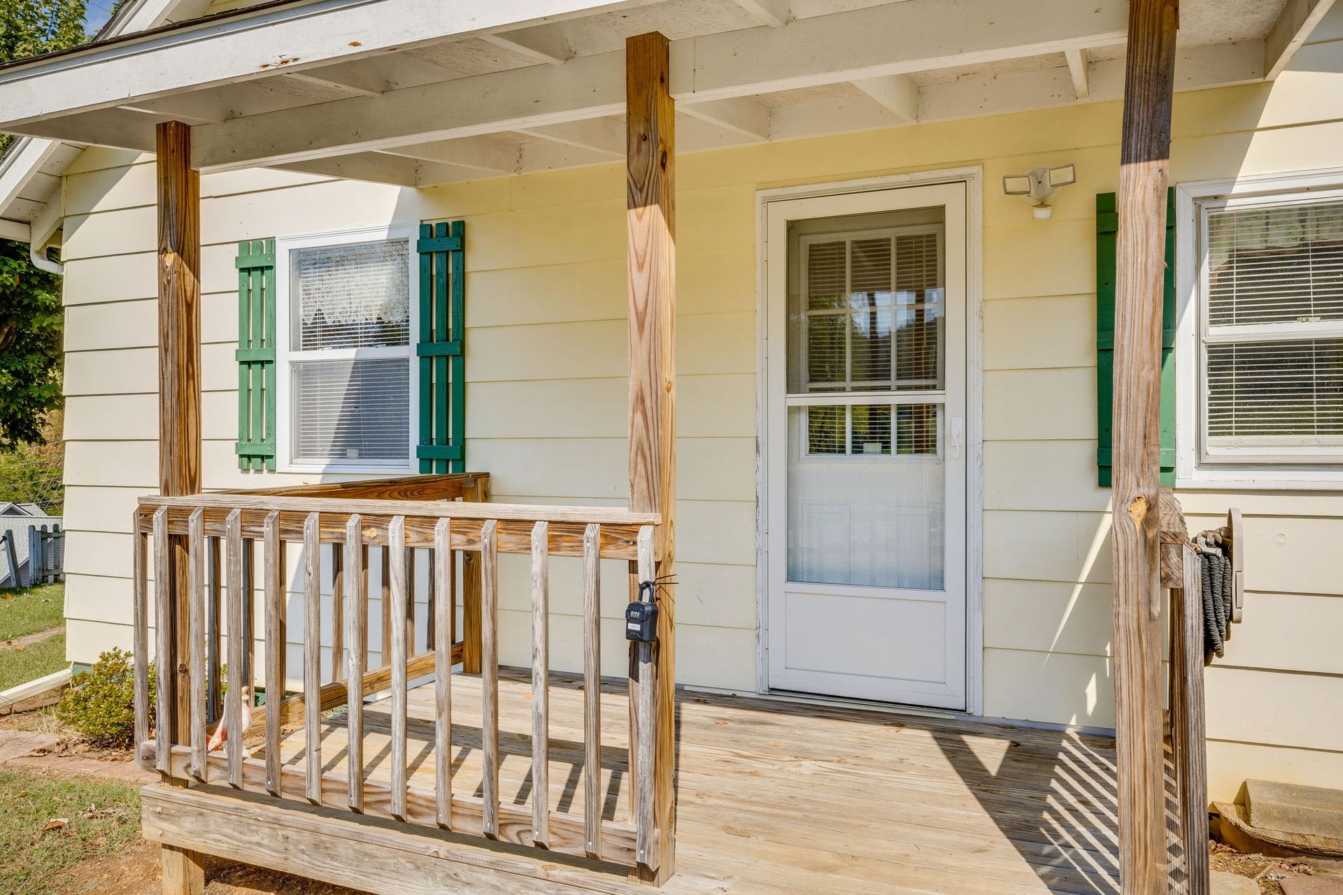 Front porch of a yellow house with a white door, green shutters, and wooden railings.