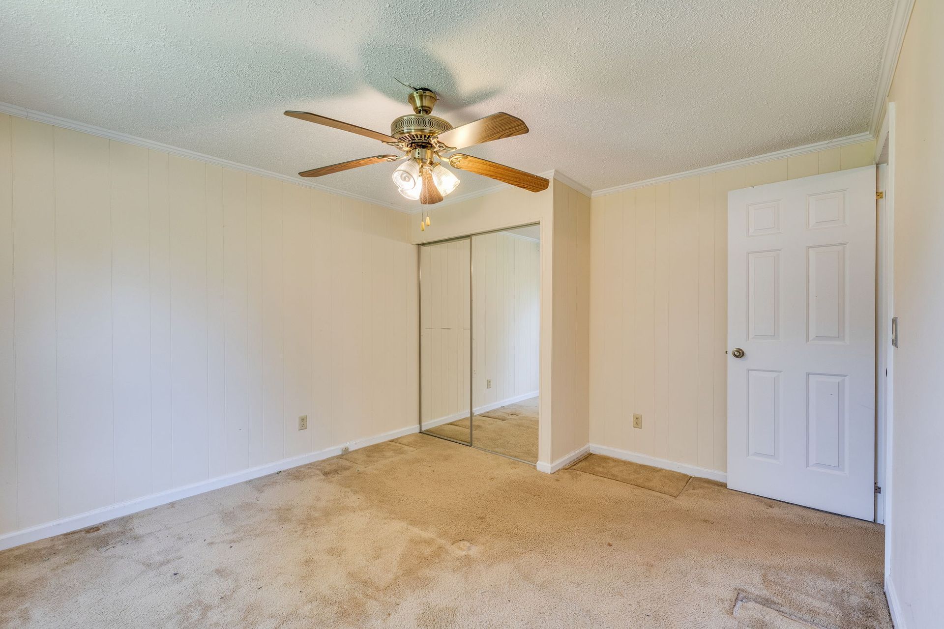 Empty beige bedroom with carpet, ceiling fan, mirrored closet, and closed white door.