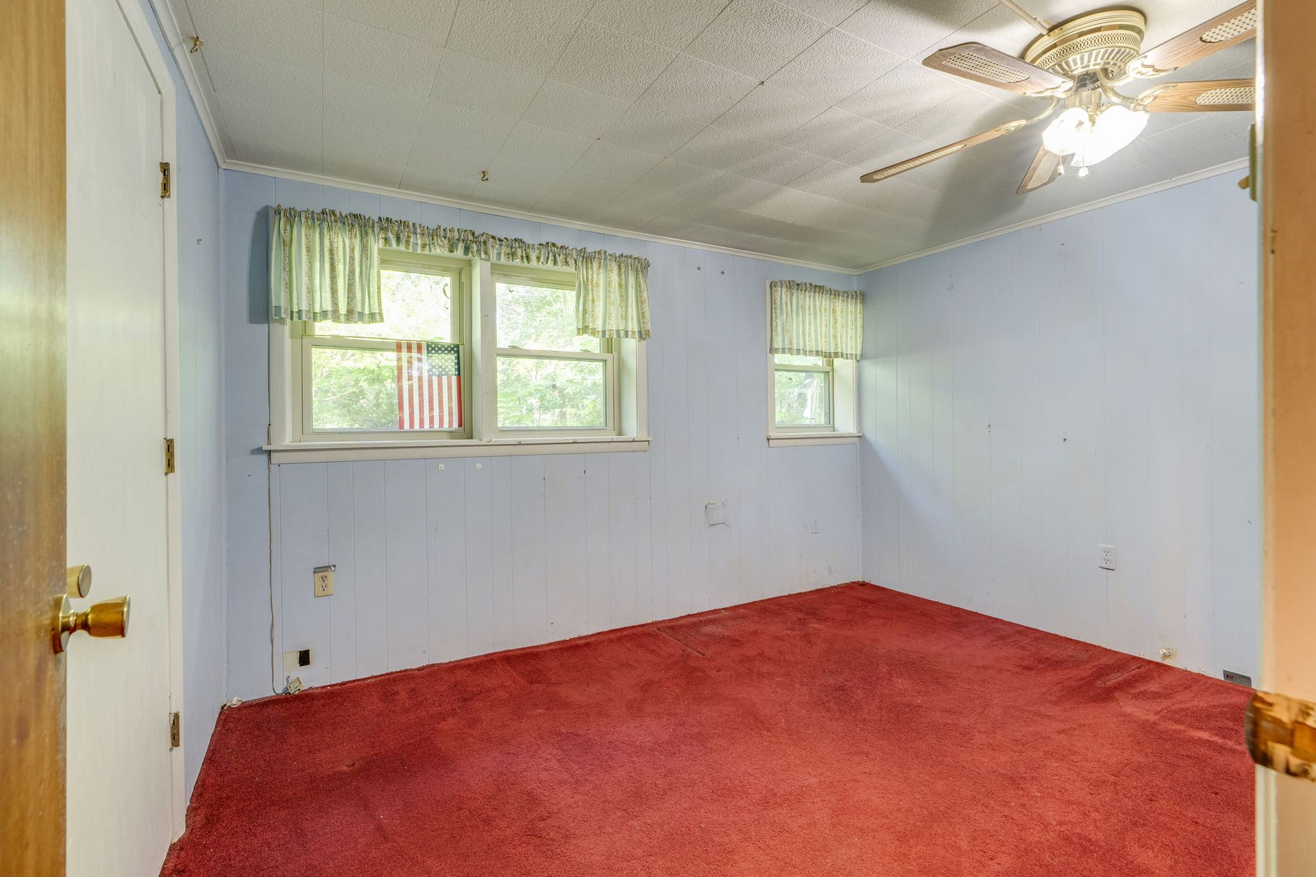 Empty room with red carpet, white walls, two windows, and a ceiling fan.