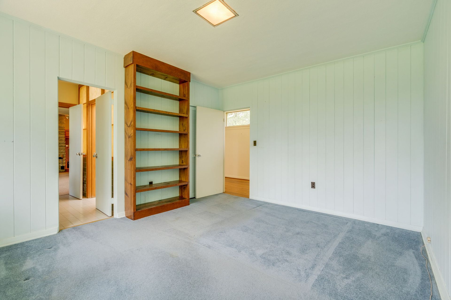 Empty carpeted room with white paneled walls and built-in wooden shelves near a doorway