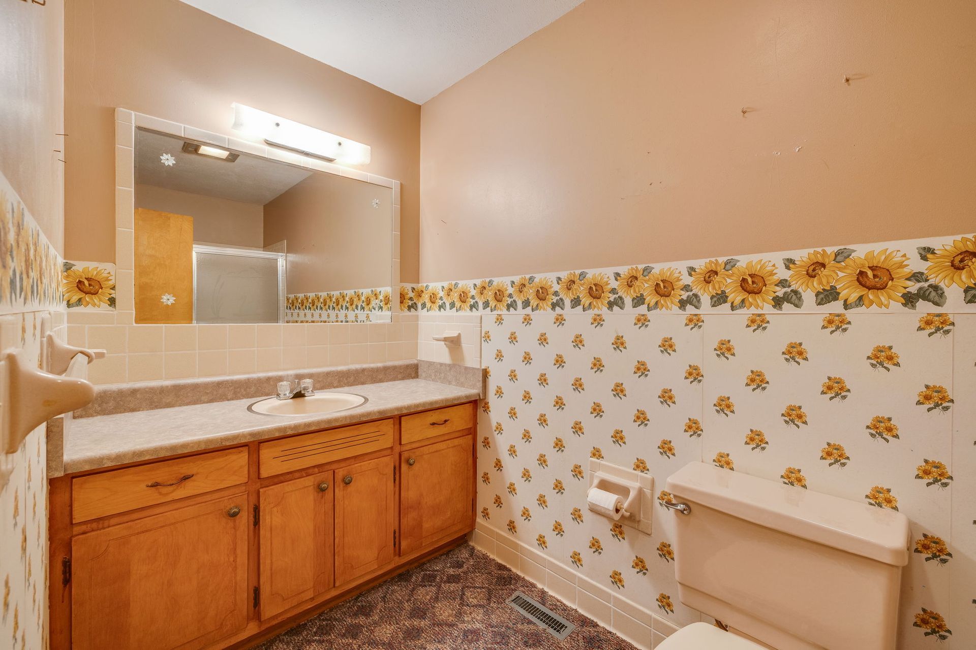 Small bathroom with wood vanity, mirror, beige walls, and yellow floral wallpaper trim.