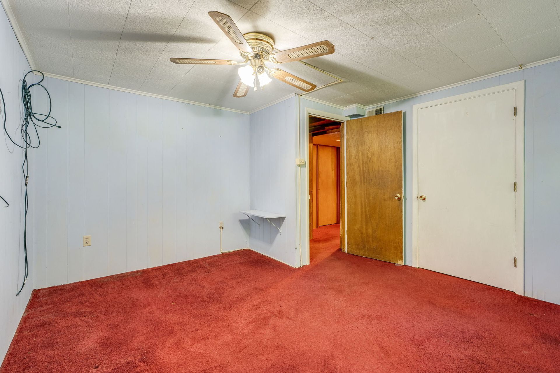 Empty bedroom with red carpet, white walls, ceiling fan, and open door to hallway