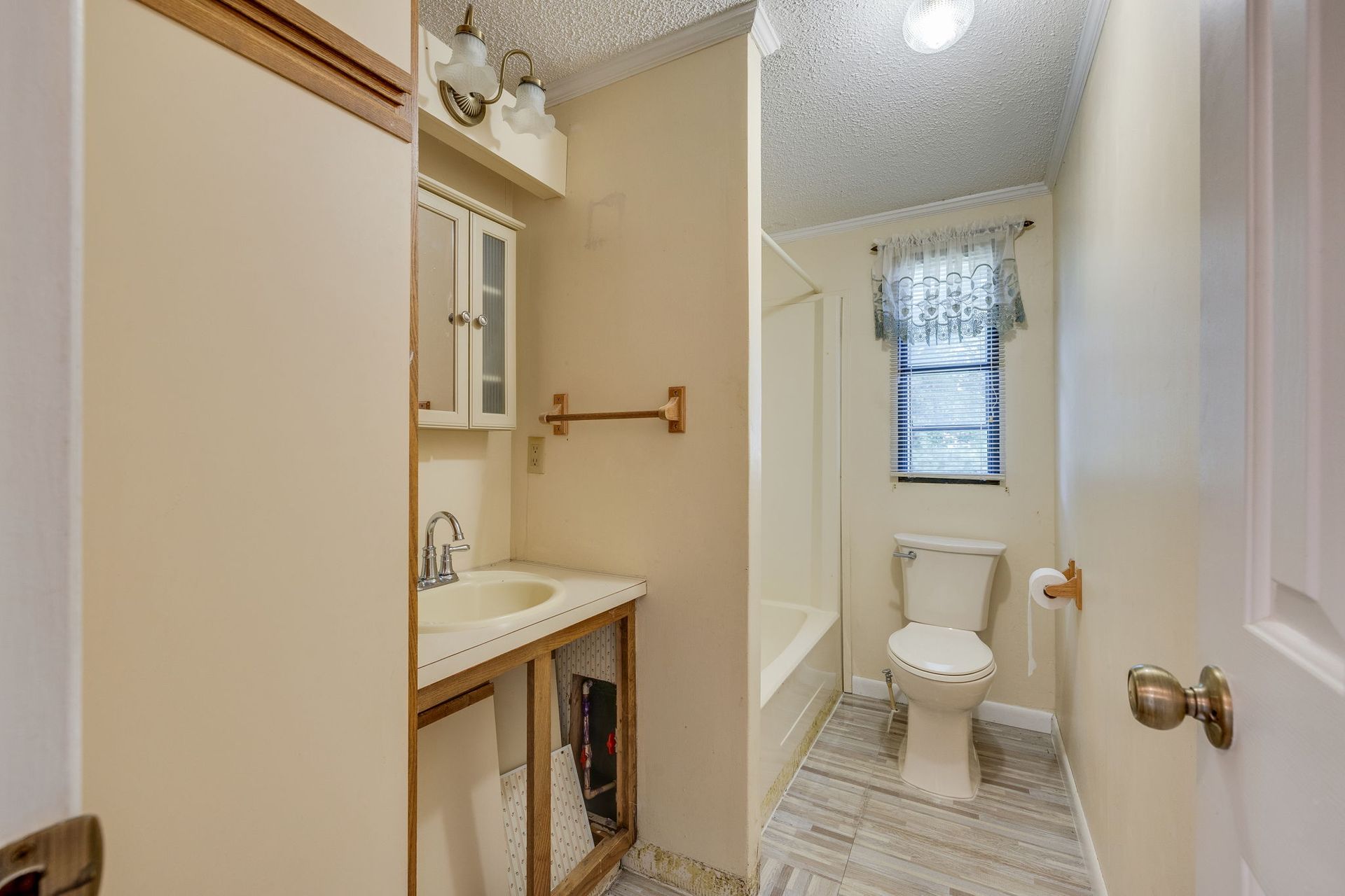 Narrow bathroom with sink, toilet, bathtub, and window with patterned curtain.