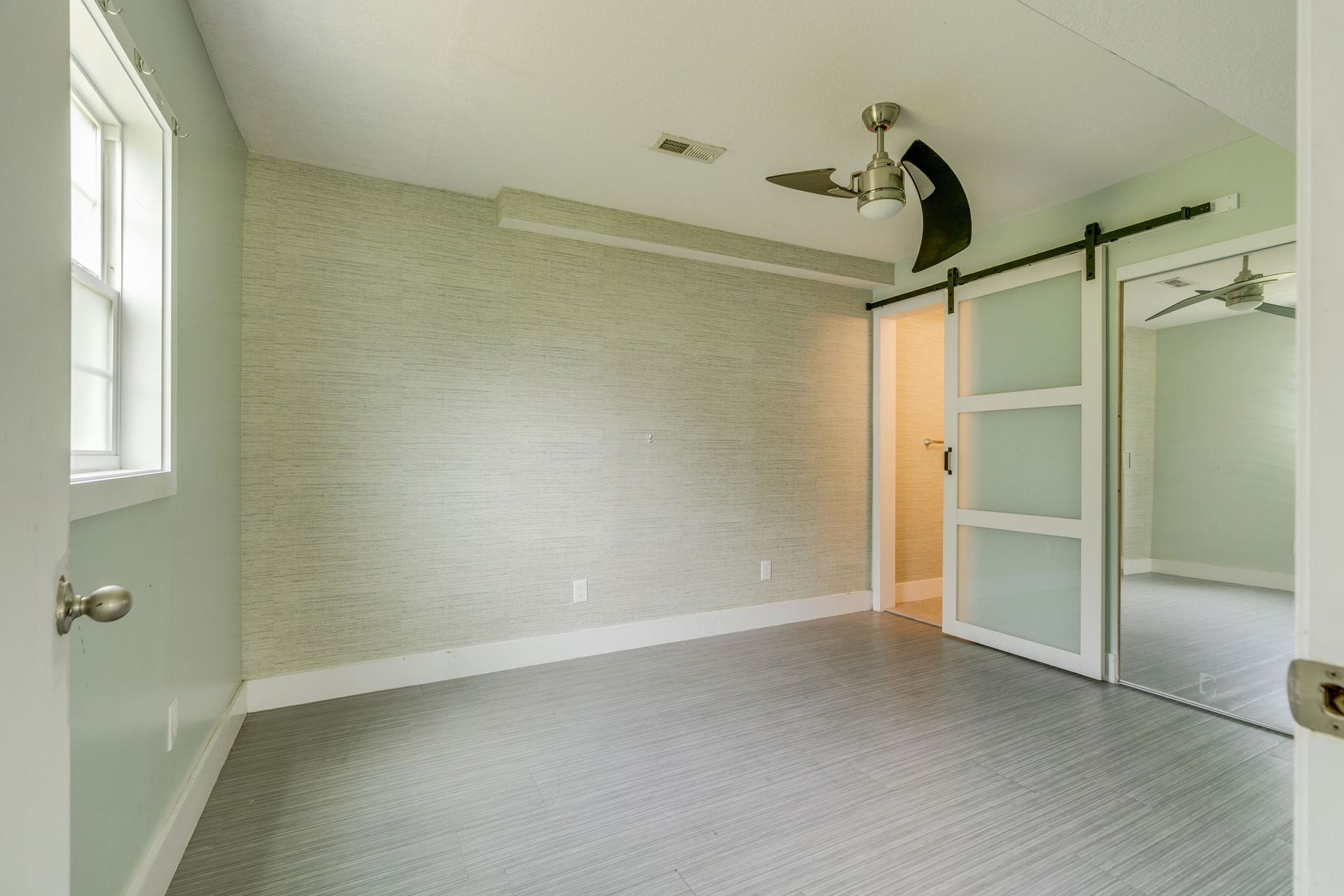 Empty bright bedroom with gray carpet, light walls, mirrored closet doors, and ceiling fan