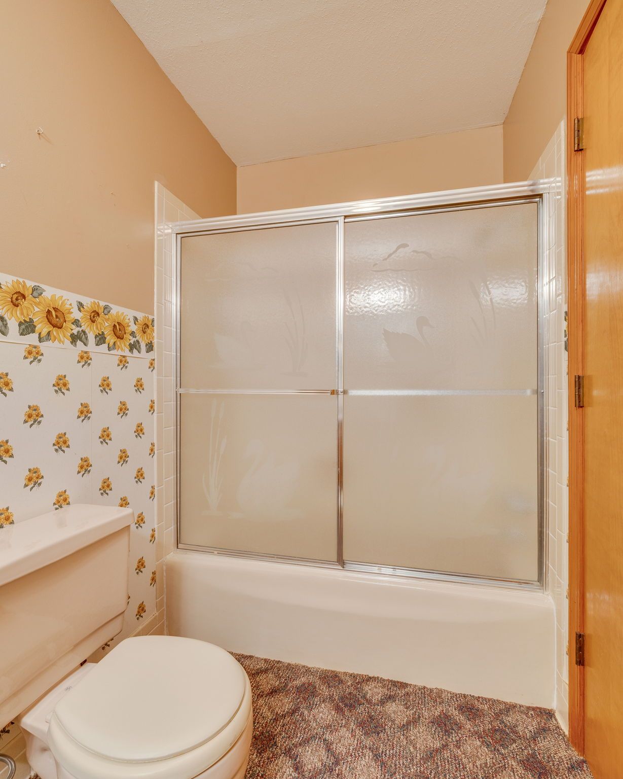 Small bathroom with tub-shower, toilet, beige walls, floral wallpaper, and wood door.