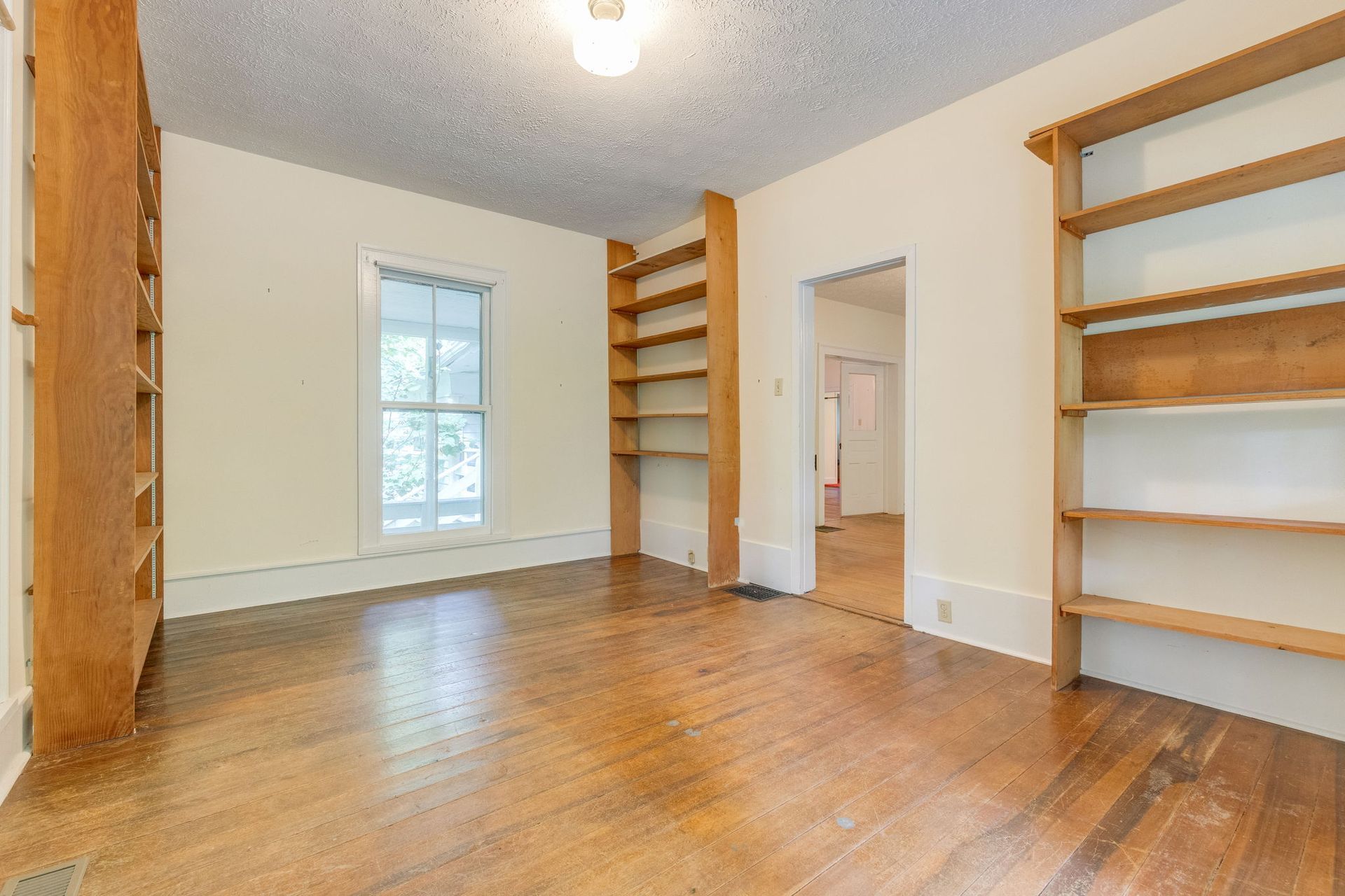 Bright empty room with hardwood floors, white walls, built-in wooden shelves, and a window