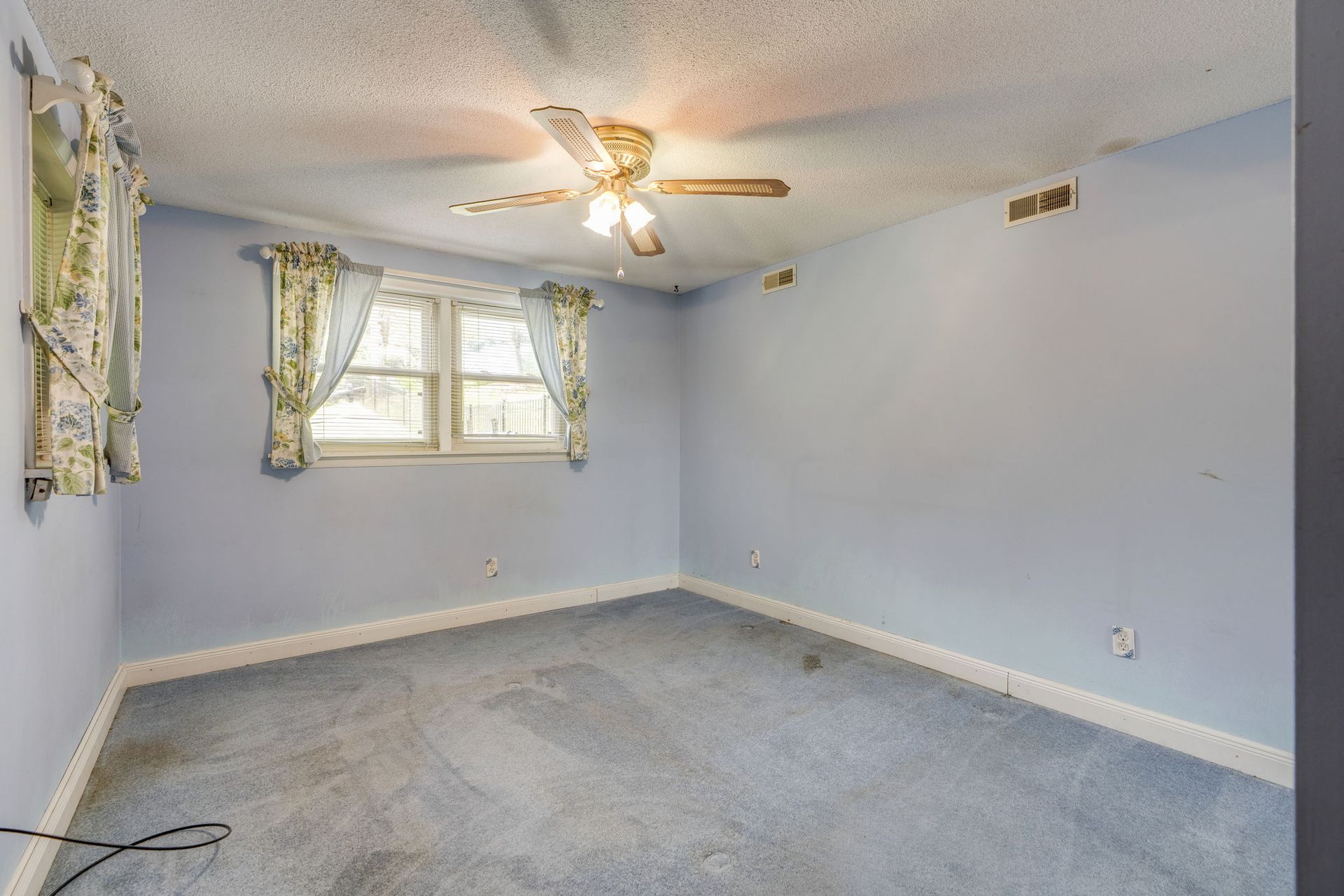 Empty light-blue bedroom with carpet, ceiling fan, window, and floral curtains