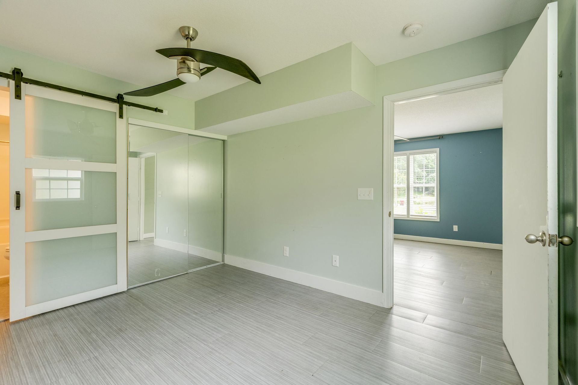 Empty mint-green bedroom with carpet, white sliding closet doors, ceiling fan, and blue adjoining room visible through doorway