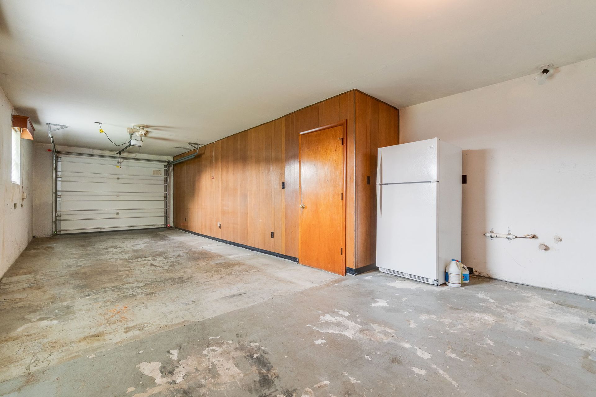 Empty garage with concrete floor, white walls, wood-paneled storage wall, and a white refrigerator.
