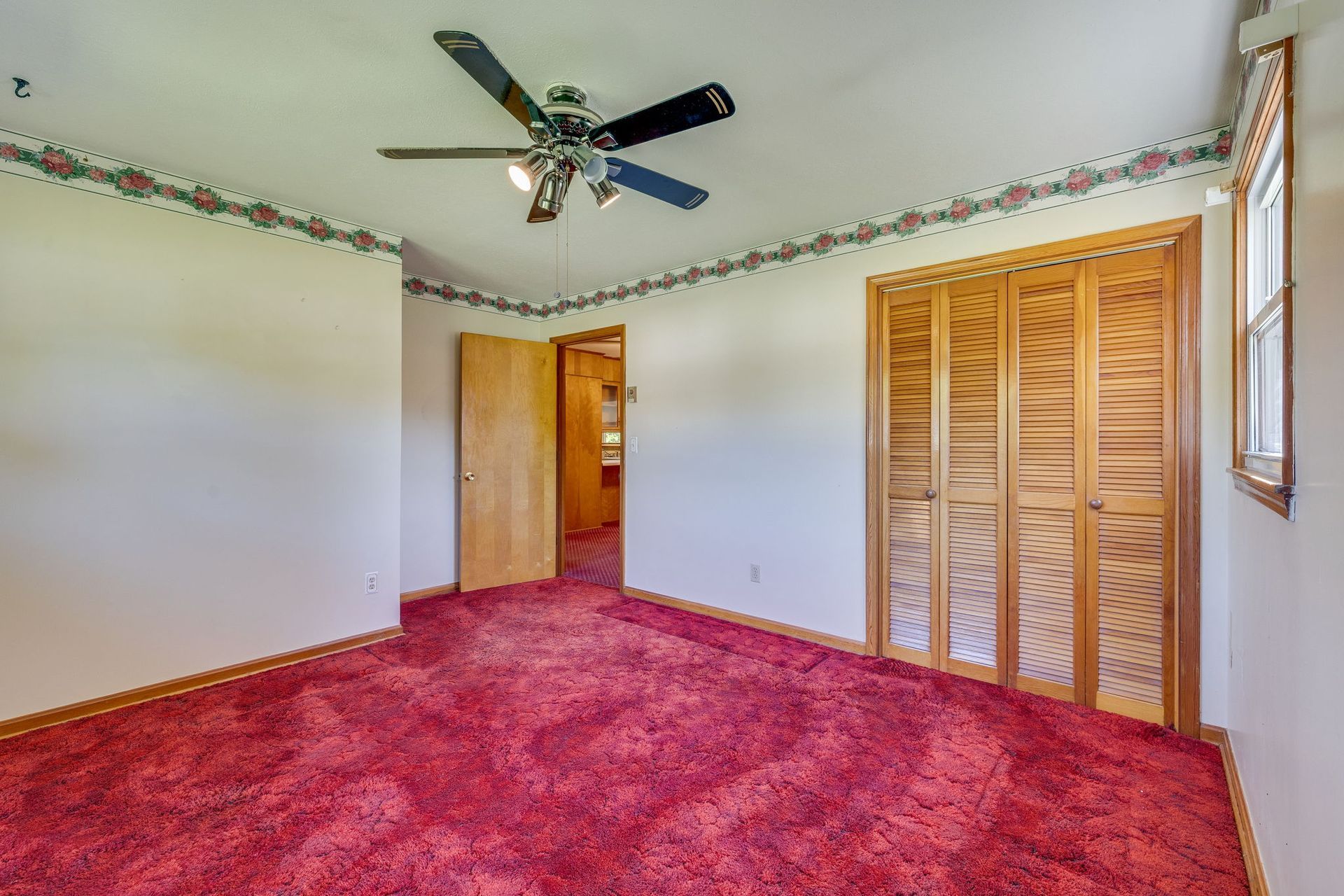 Empty bedroom with red carpet, white walls, ceiling fan, and wood closet doors