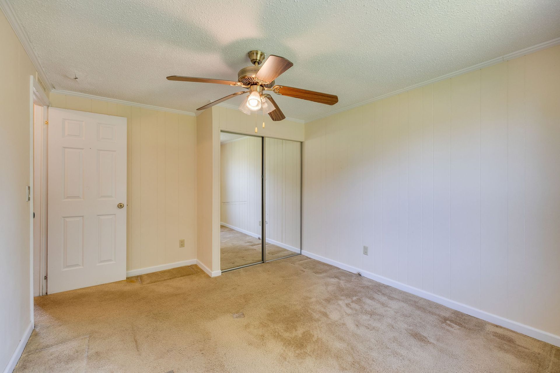 Empty beige bedroom with carpet, mirrored closet doors, white walls, and a ceiling fan