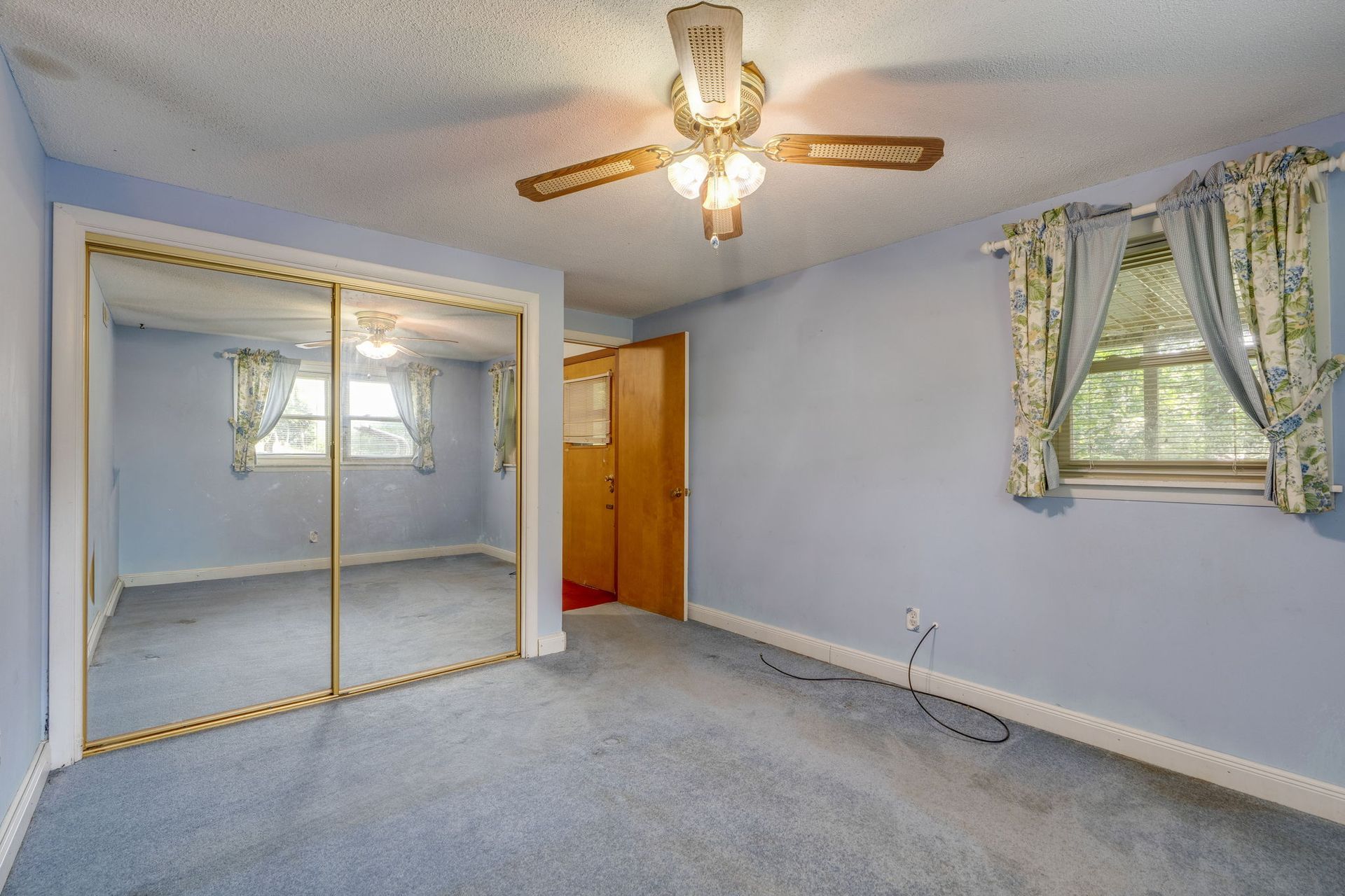 Empty light-blue bedroom with carpet, mirrored closet doors, ceiling fan, and window with curtains.