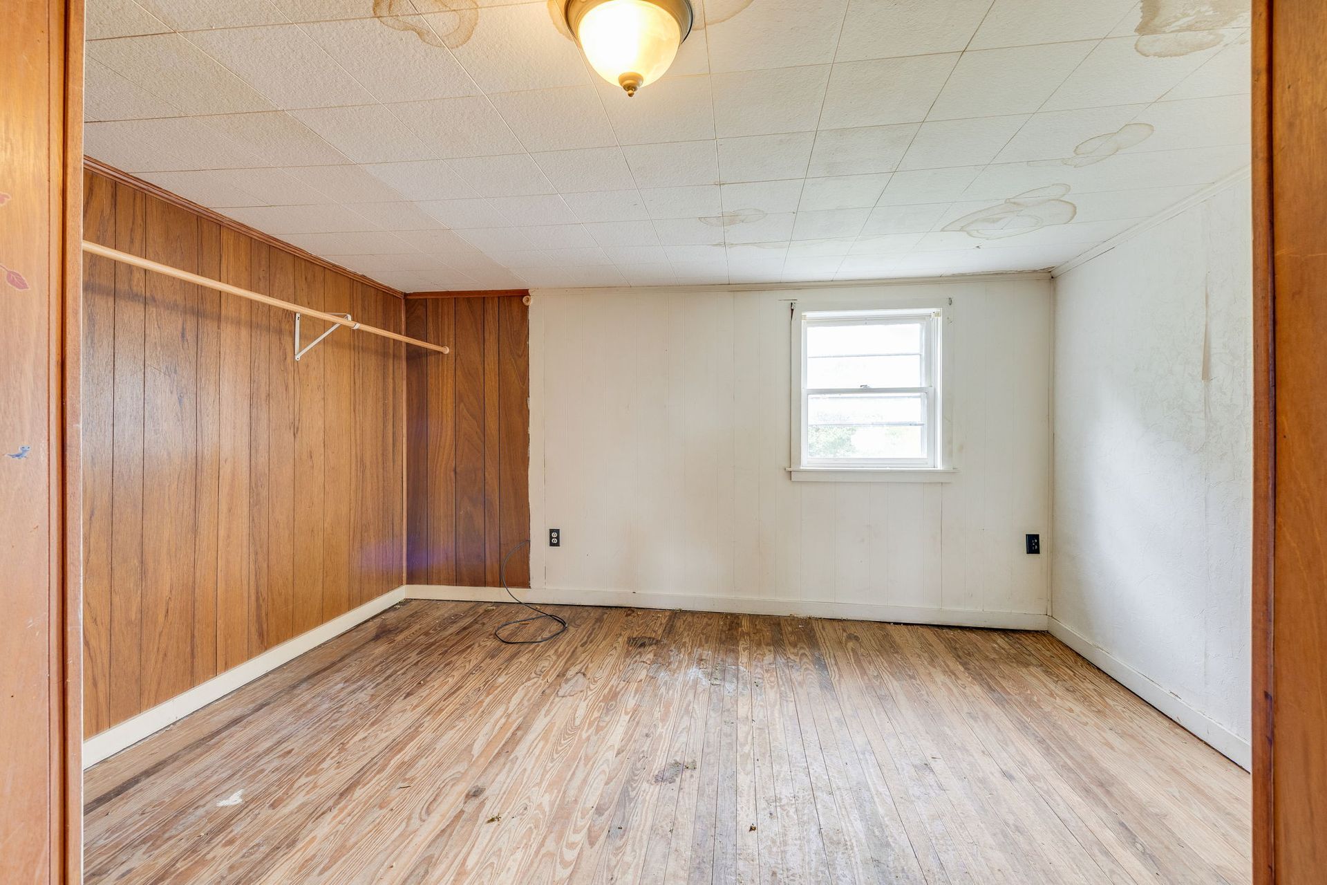 Empty room with wood-paneled wall, white walls, worn hardwood floor, and a small window.