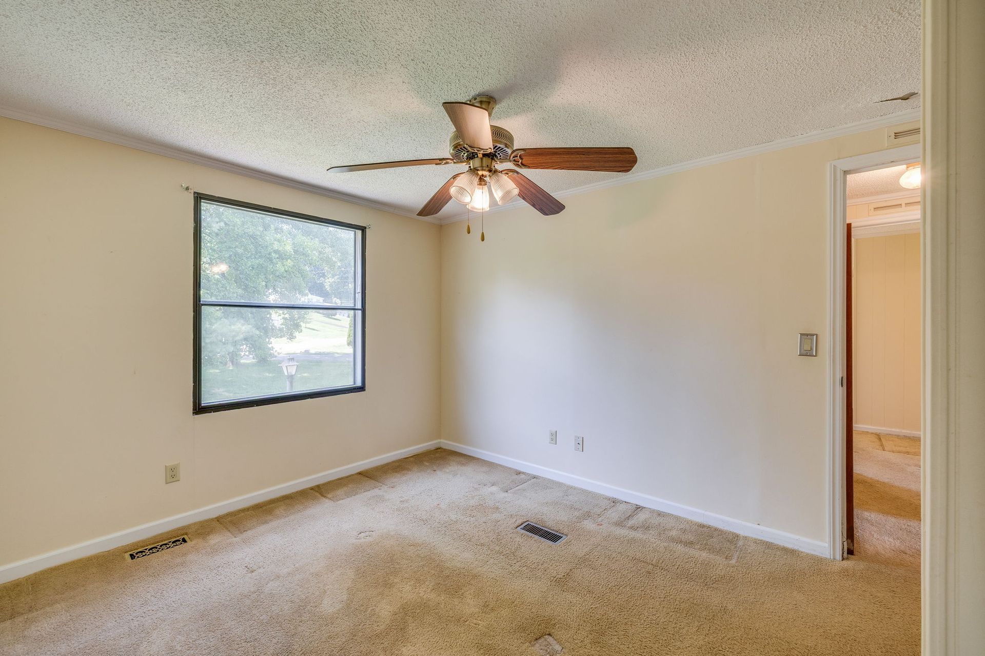Empty beige carpeted bedroom with window, ceiling fan, and open doorway