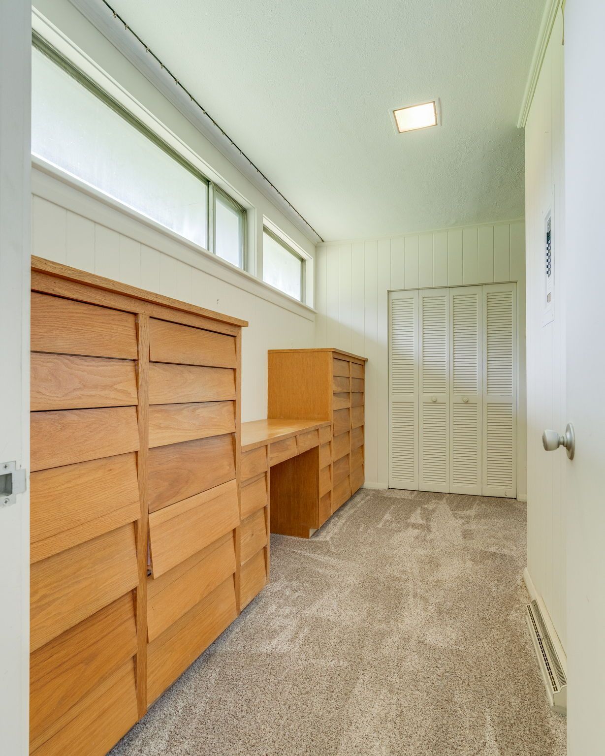 Bright walk-in closet with wooden shelves, louvered doors, high windows, and carpeted floor