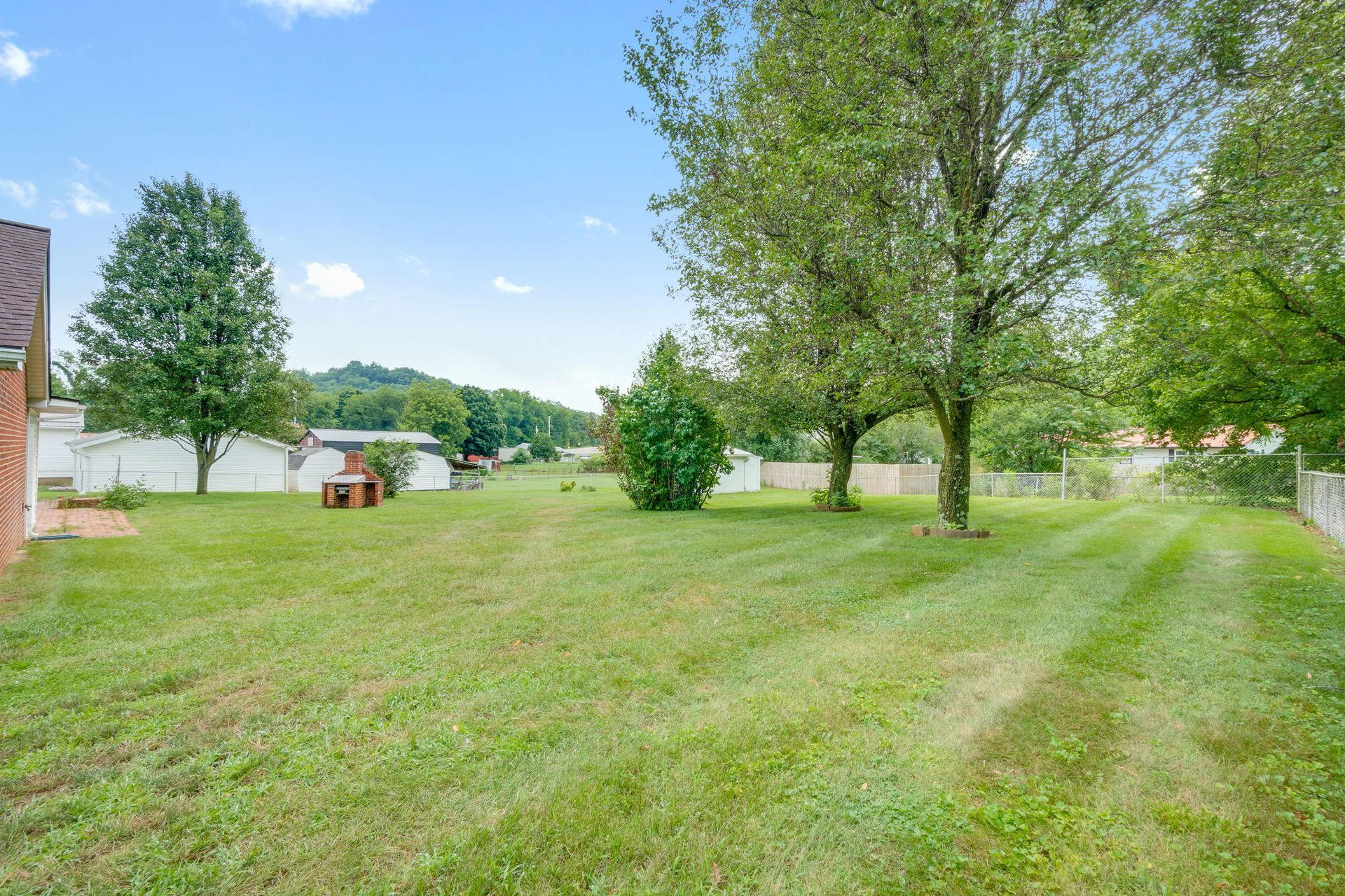 Grassy backyard with mature trees, a small shed, and a distant view of neighboring homes under a blue sky