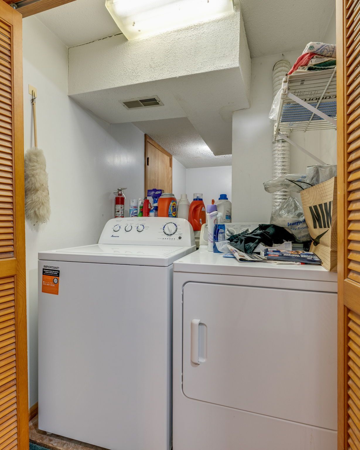 Laundry room with white washer and dryer under a sloped ceiling, shelves of cleaning supplies, and a broom.