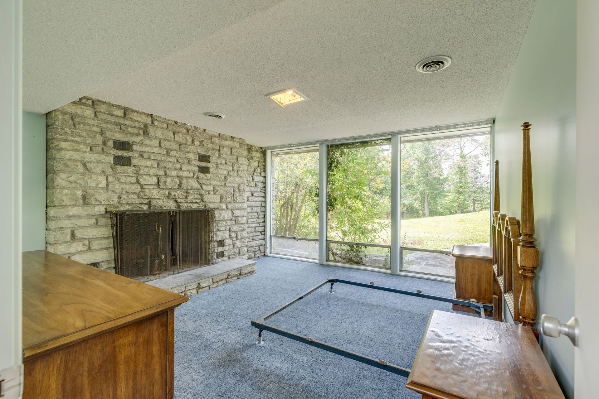 Empty living room with stone fireplace, large windows, carpet, and a wooden dresser near the wall