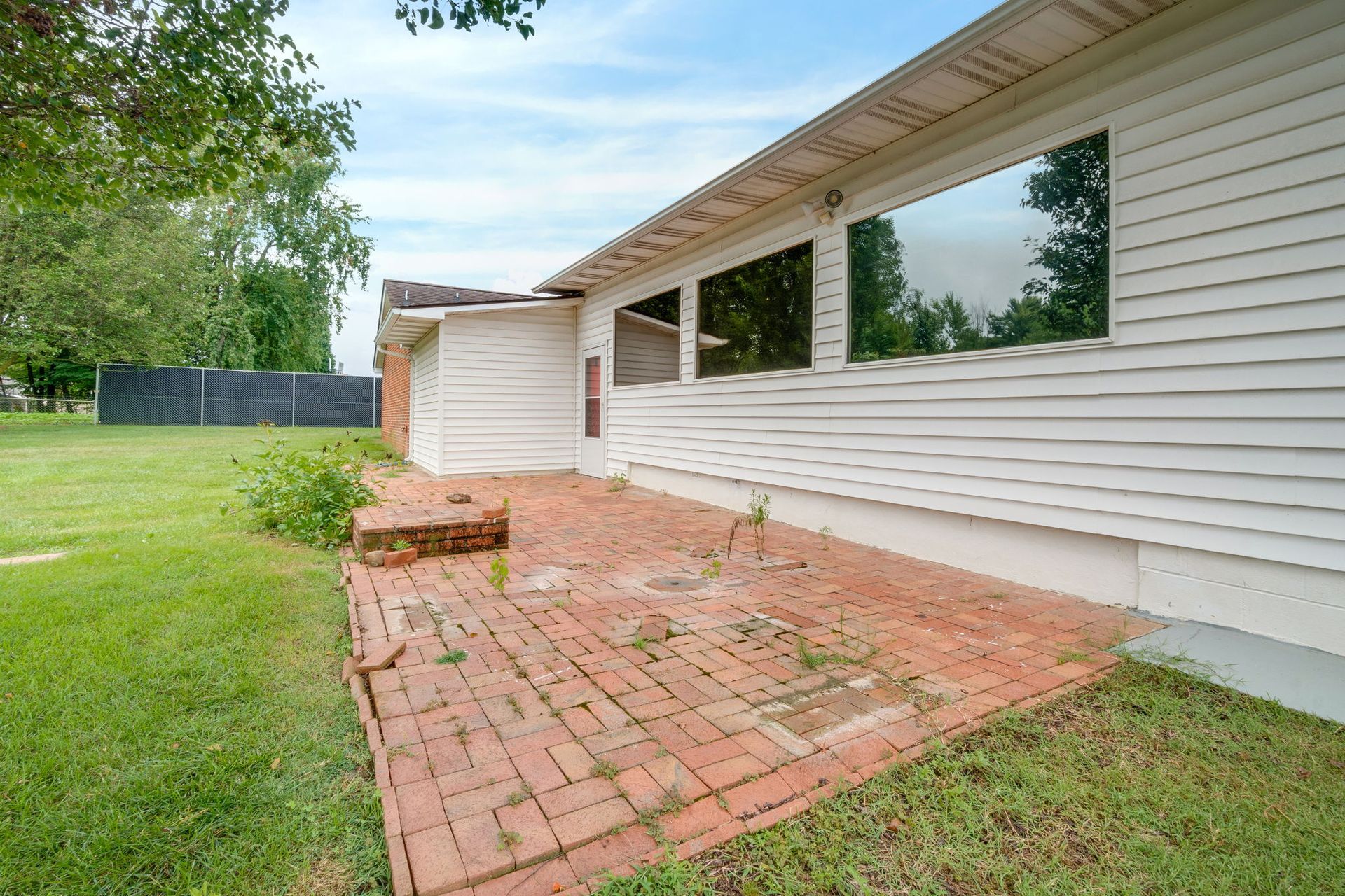 Brick patio beside white house with a window, green lawn, and trees under an overcast sky