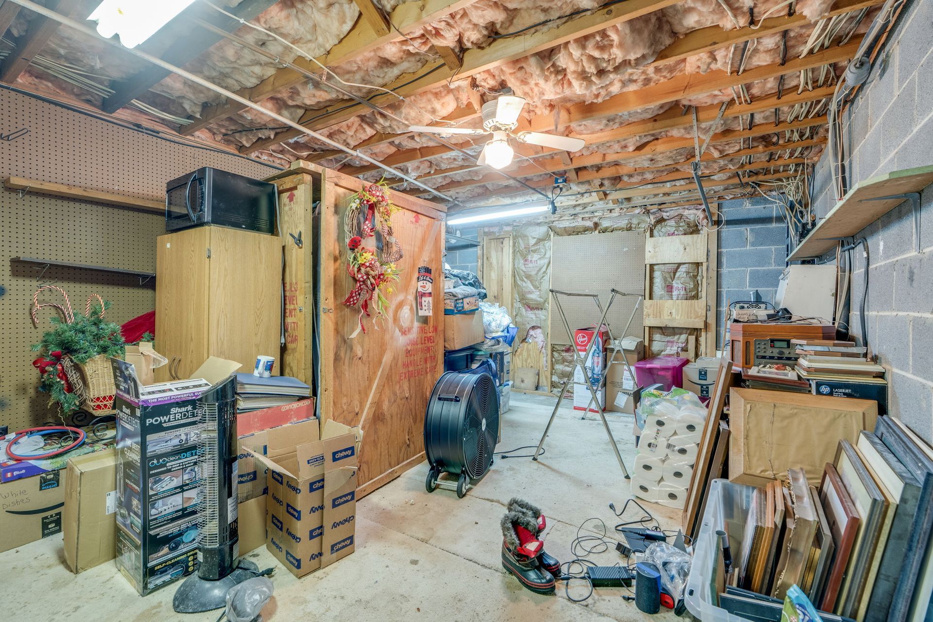 Cluttered basement workshop with exposed ceiling joists, tools, wood panels, and stacked boxes.