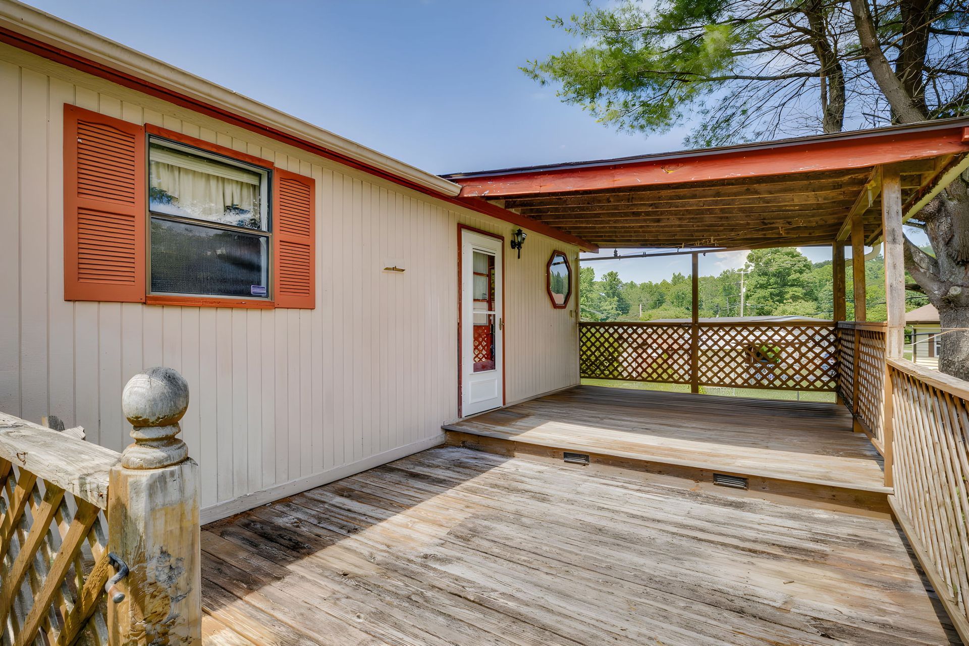 Front porch with weathered wooden deck, beige siding, red trim, and a covered seating area overlooking trees