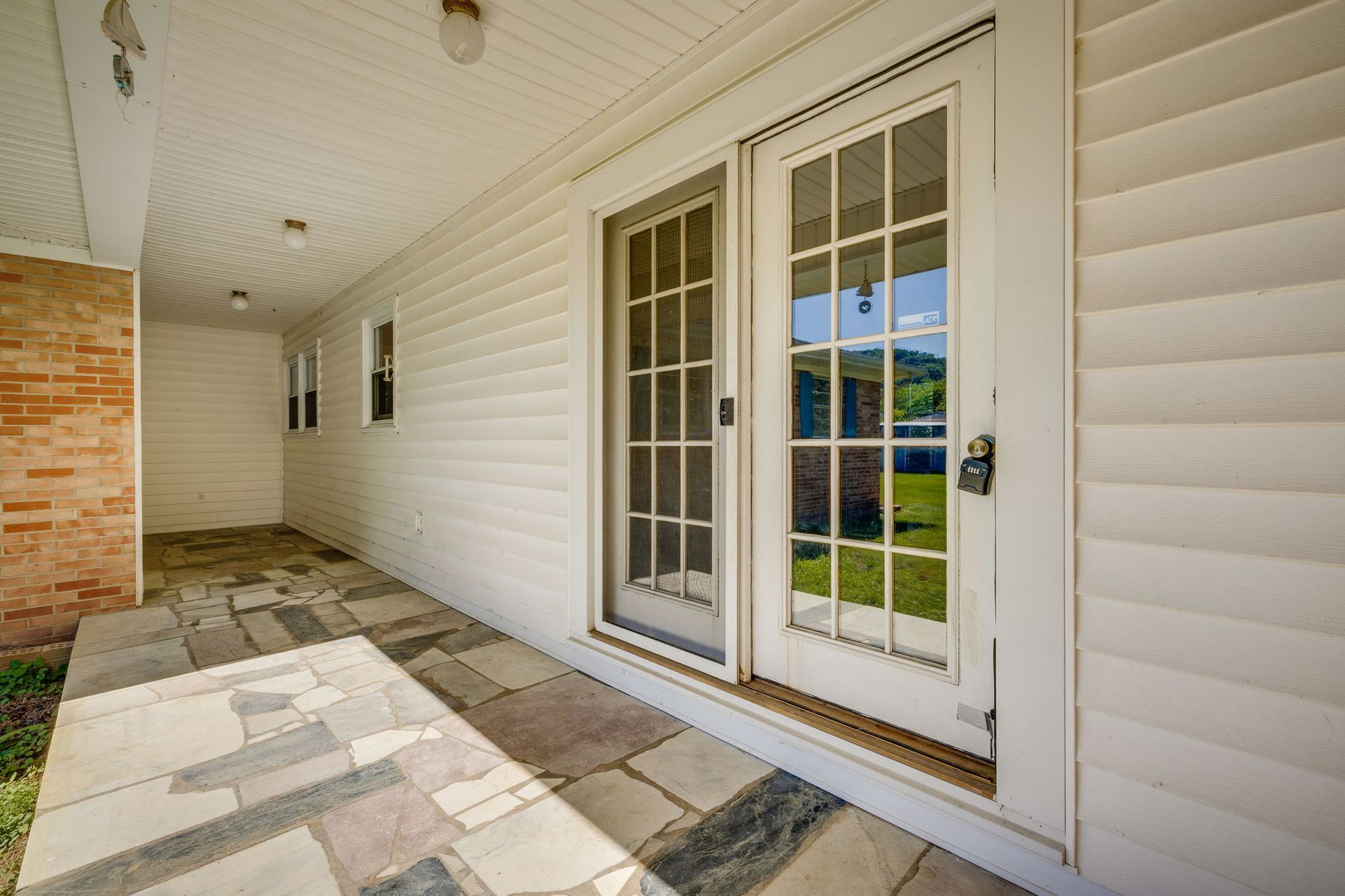 Covered porch with white siding, French doors, and a stone-tiled floor beside a brick wall.